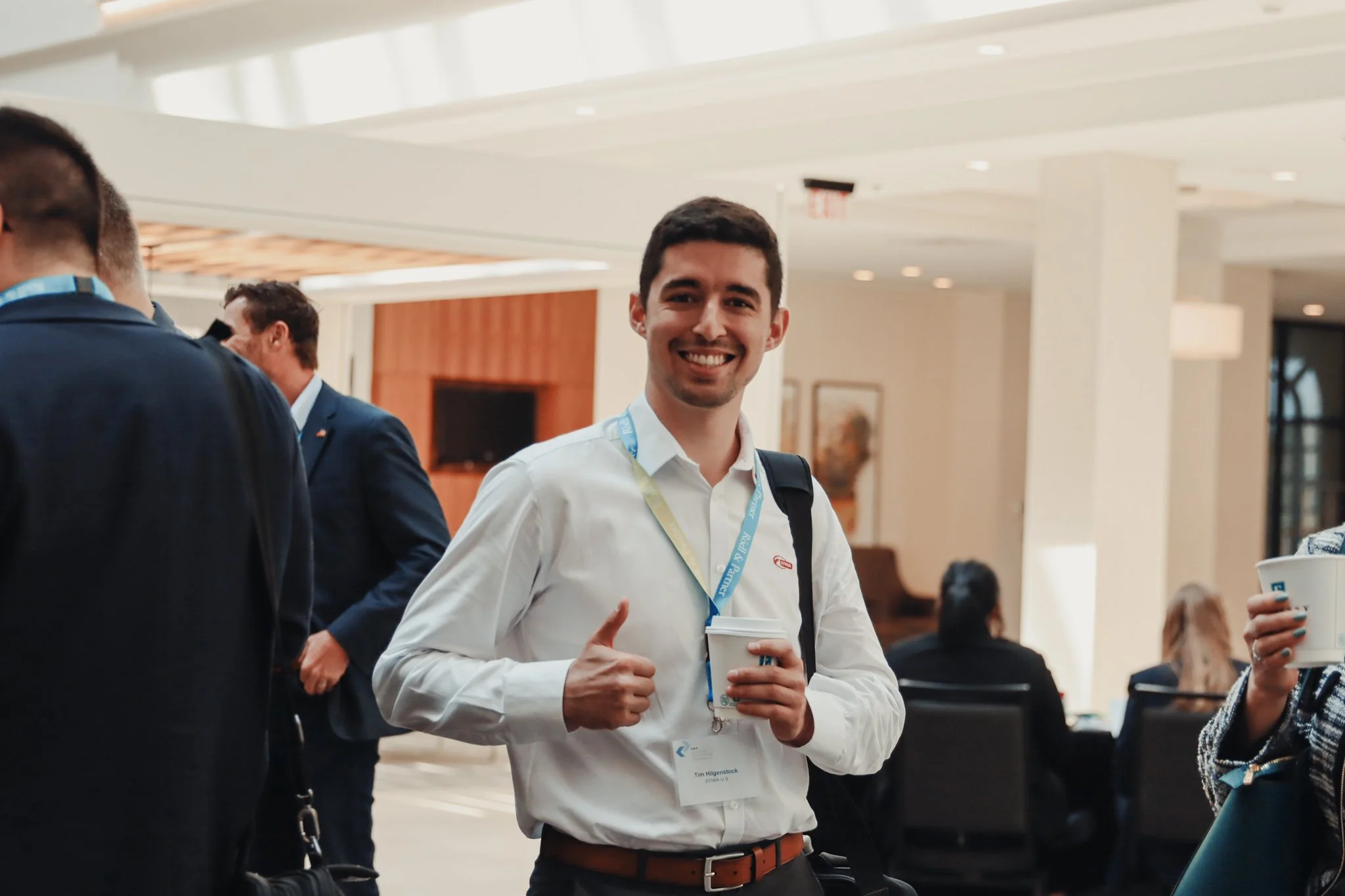 Young man in white shirt smiling at a professional conference or networking event, holding a coffee cup, with other attendees in the background.