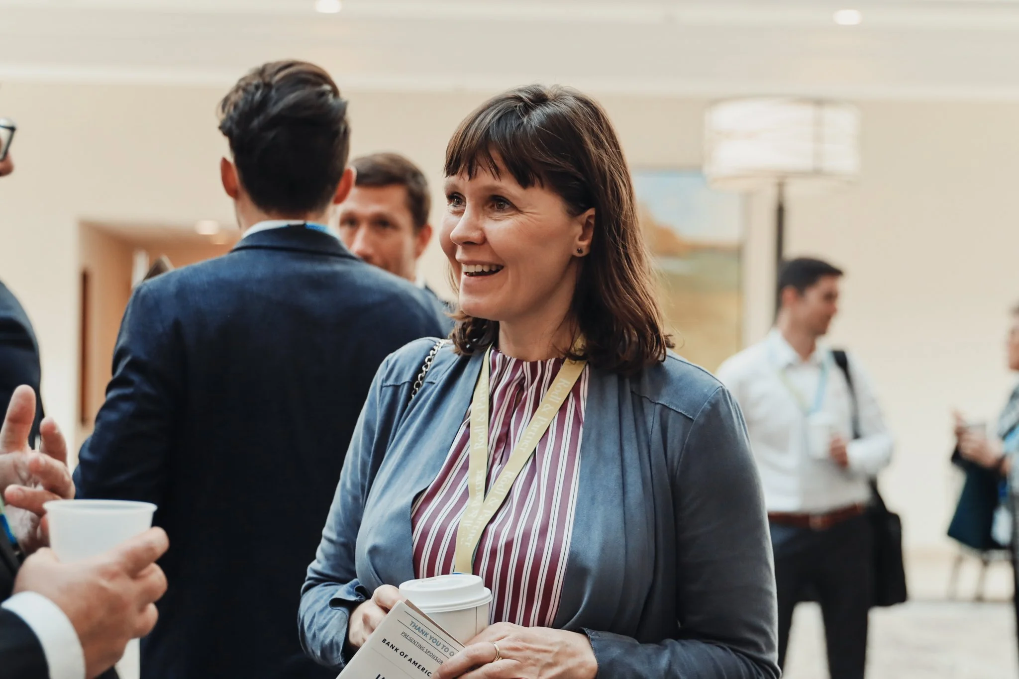A woman with short brown hair, wearing a blue blazer and striped shirt, is smiling and holding a coffee cup and a paper, while talking to a man at what appears to be a conference or networking event. Several people are in the background, and the sett