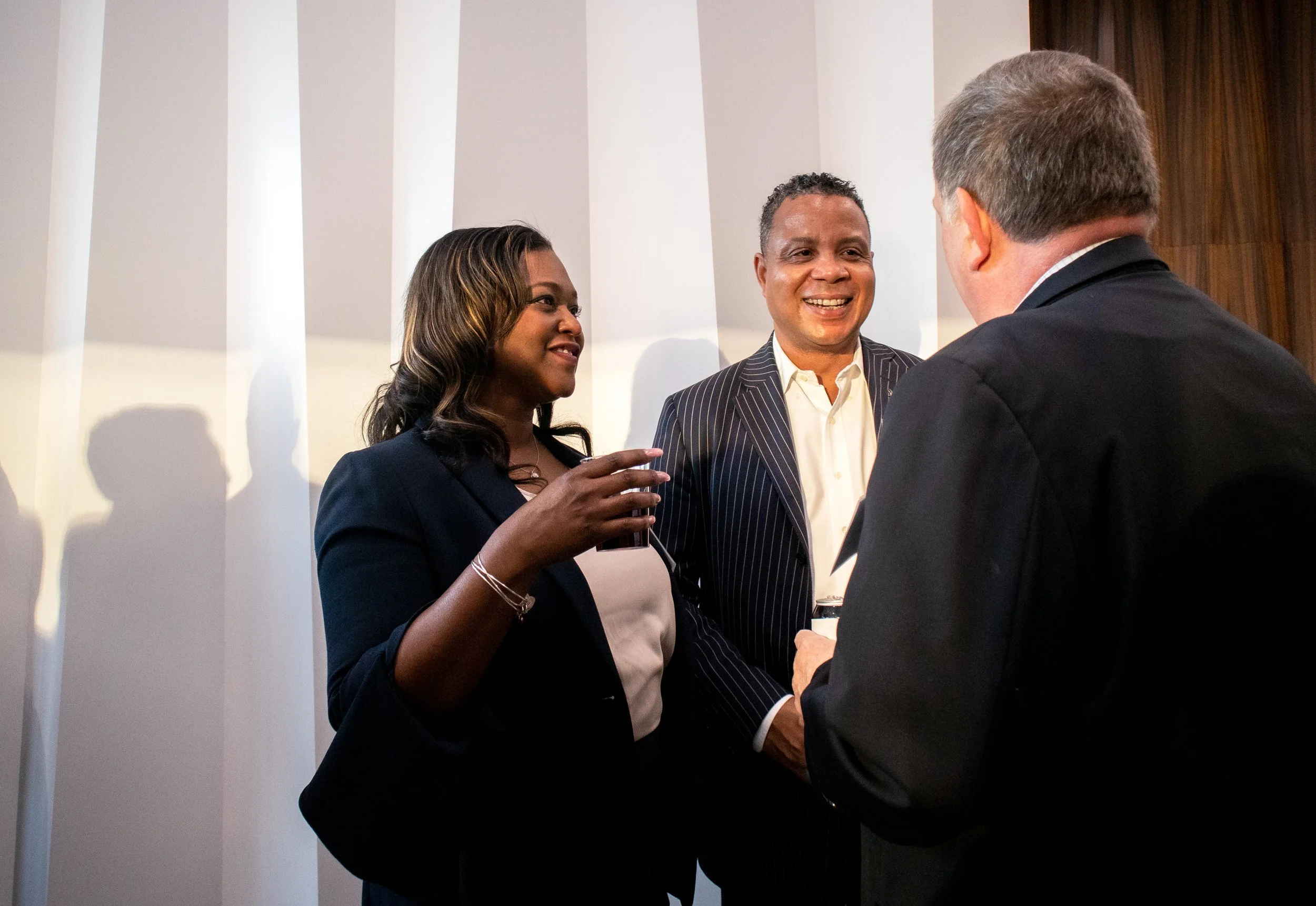 Three people are engaged in a conversation in a well-lit room. The woman on the left has dark, wavy hair and is wearing a navy blazer. The man in the middle has short gray hair, is smiling, and is wearing a black pinstripe suit with a white shirt. Th