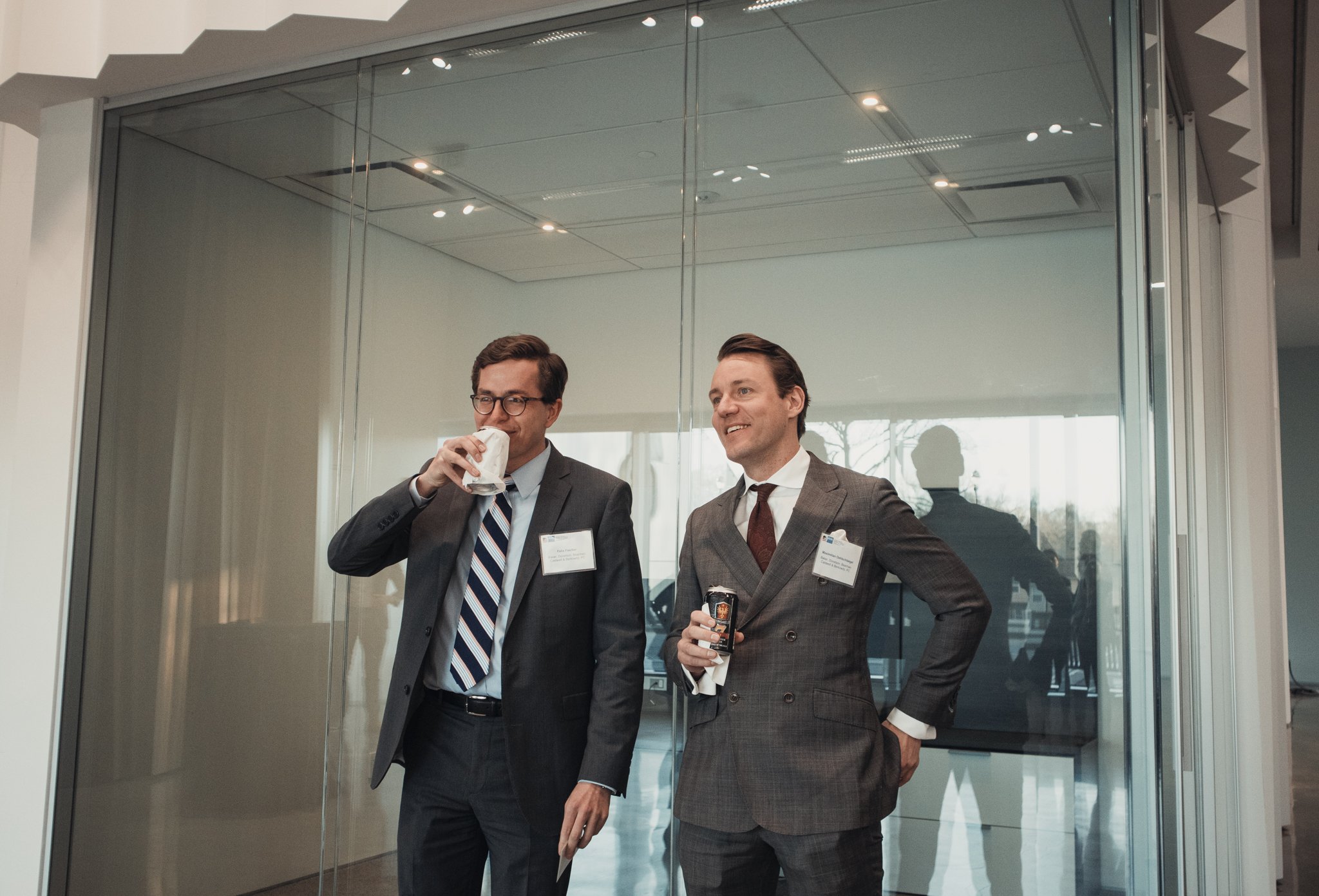 Two men in business suits standing together near glass elevator doors in a modern building, smiling and holding drinks.