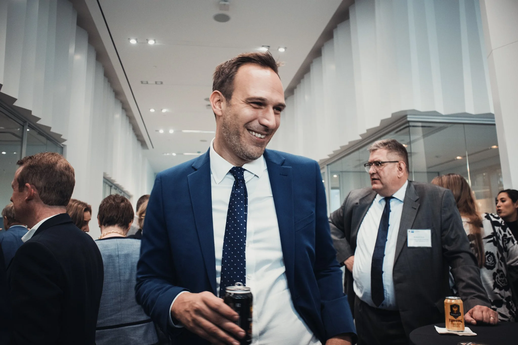 A man in a blue suit with a white shirt and blue polka dot tie is smiling while holding a drink at a crowded social gathering or conference in a modern building.
