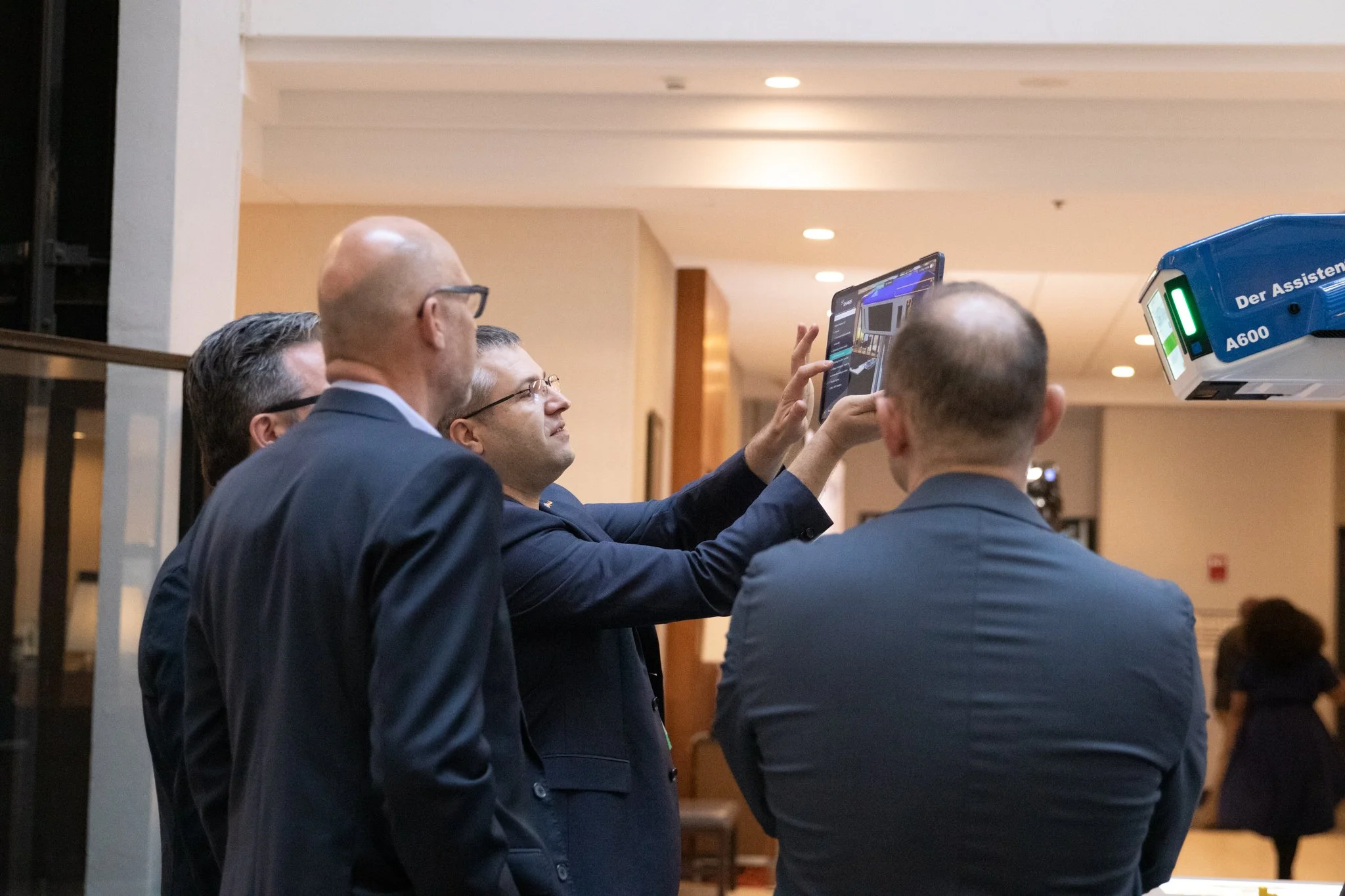 Group of professionally dressed men in suits gathered around a tablet computer, engaging in discussion at an indoor event.