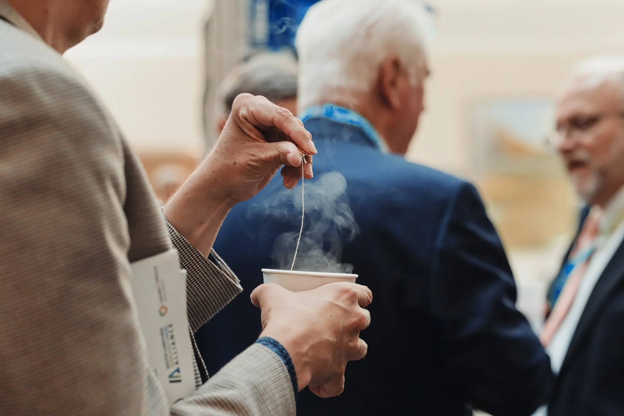 People sitting in a row, one person holding a coffee cup and stirring, others speaking or listening in an indoor setting.