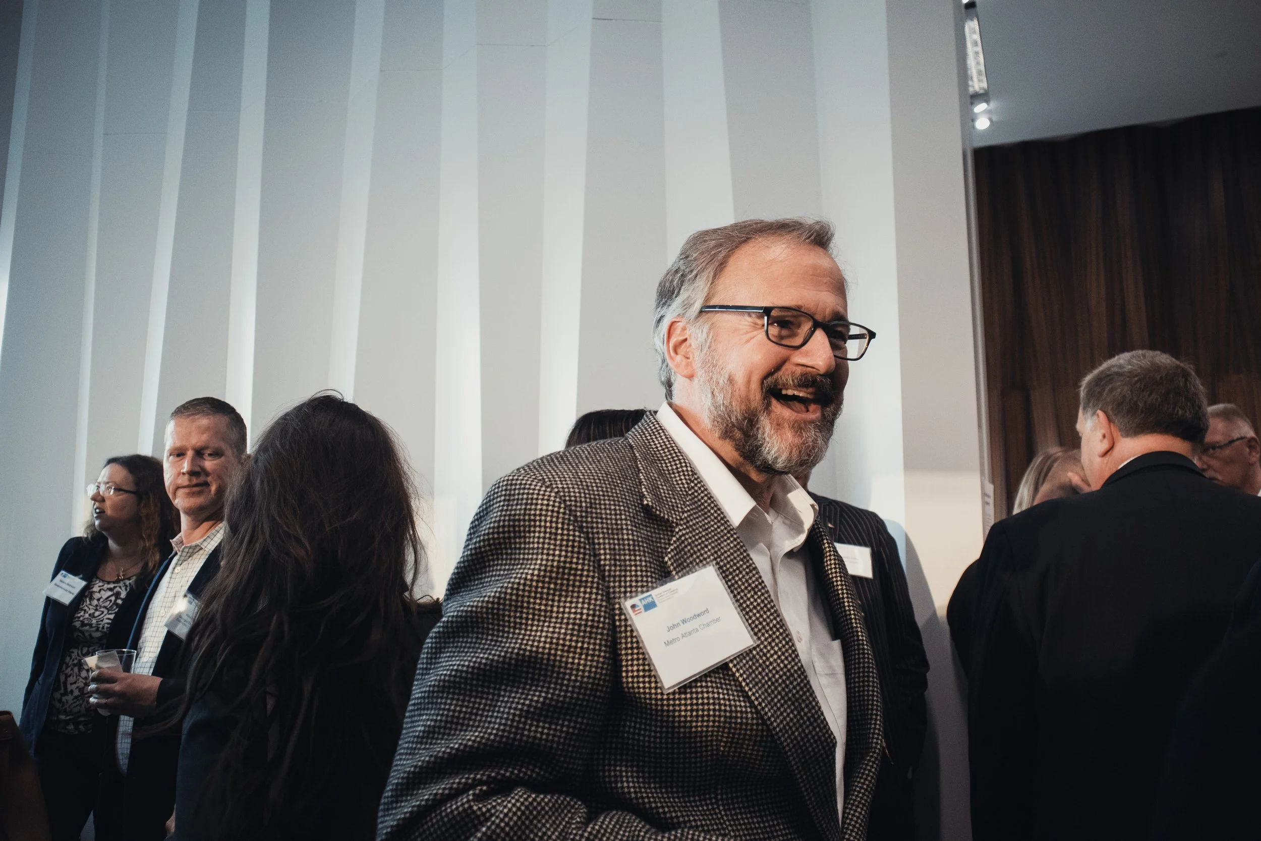 Man with glasses smiling at event, surrounded by other people in a modern conference room with white and dark wood wall panels.