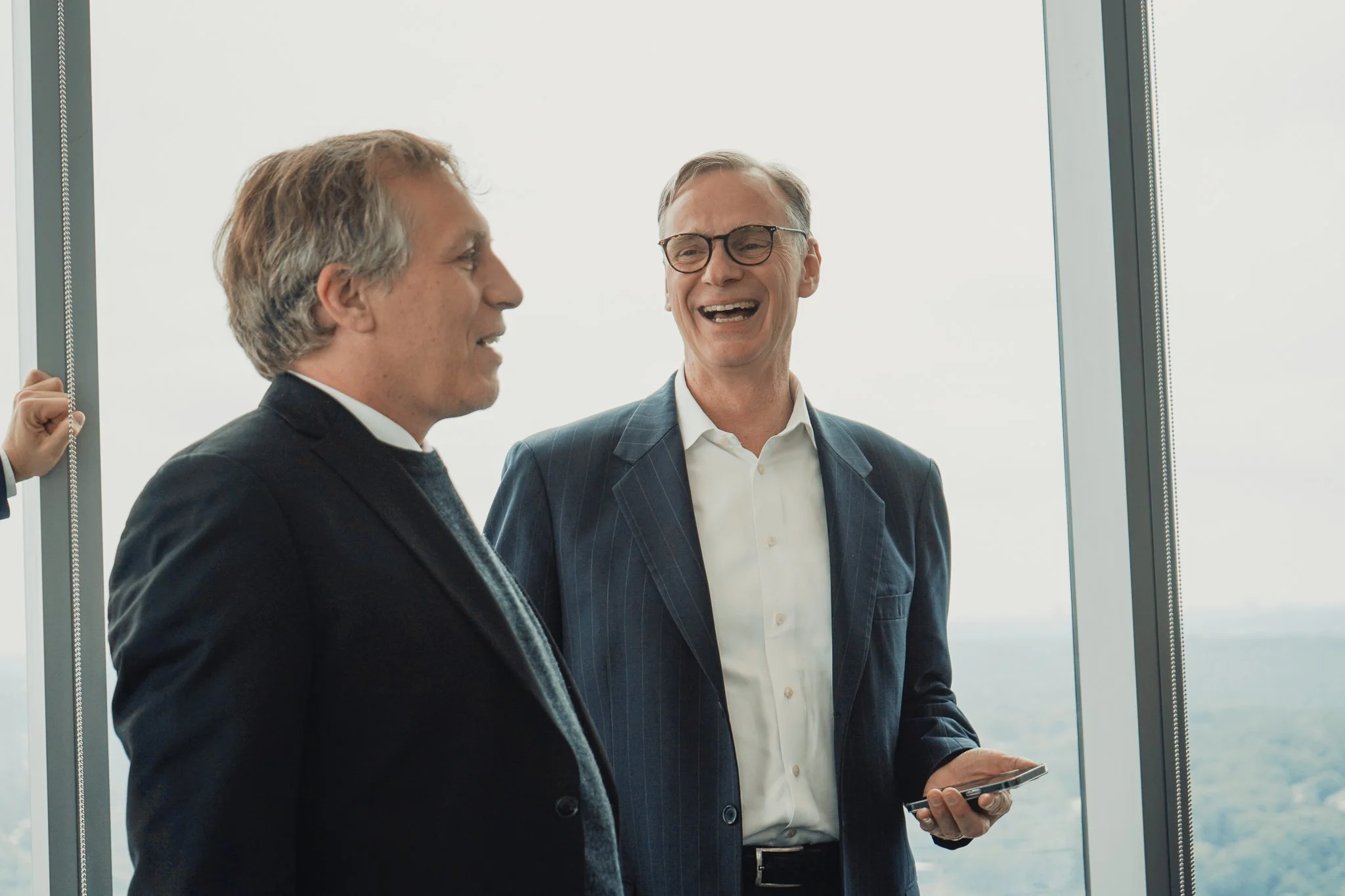 Two men in business suits smiling and talking, one holding a smartphone, inside an office building with large windows showing an outdoor view.