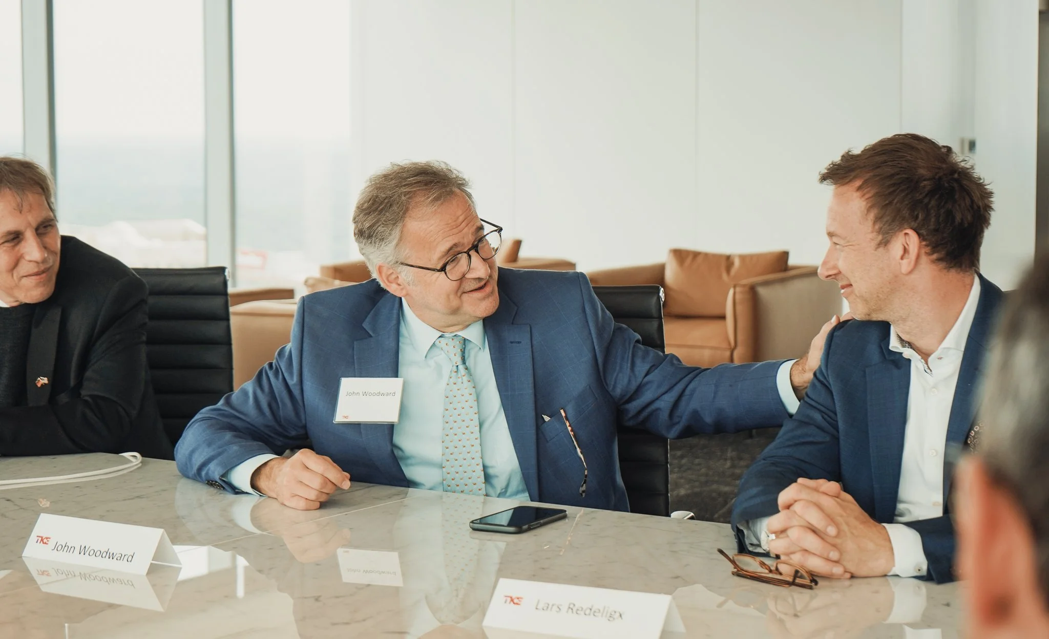 Three men in business attire sitting at a conference table, smiling and engaging in conversation during a professional meeting, with name tags visible.