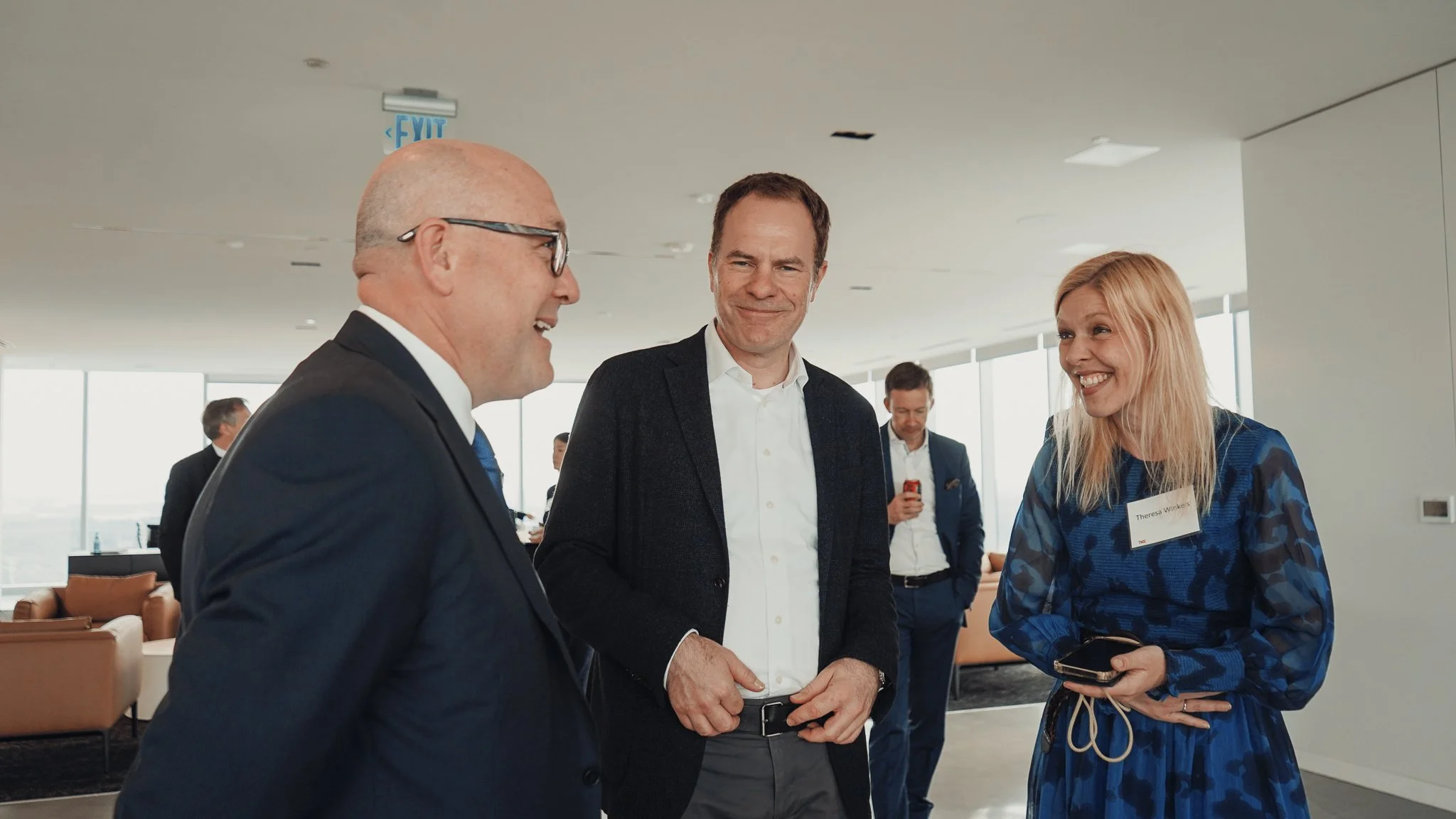 Three people are standing together in a modern indoor space, engaged in conversation. One is a bald man with glasses wearing a dark suit, another is a man with short hair wearing a dark blazer and white shirt, and the woman has shoulder-length blonde