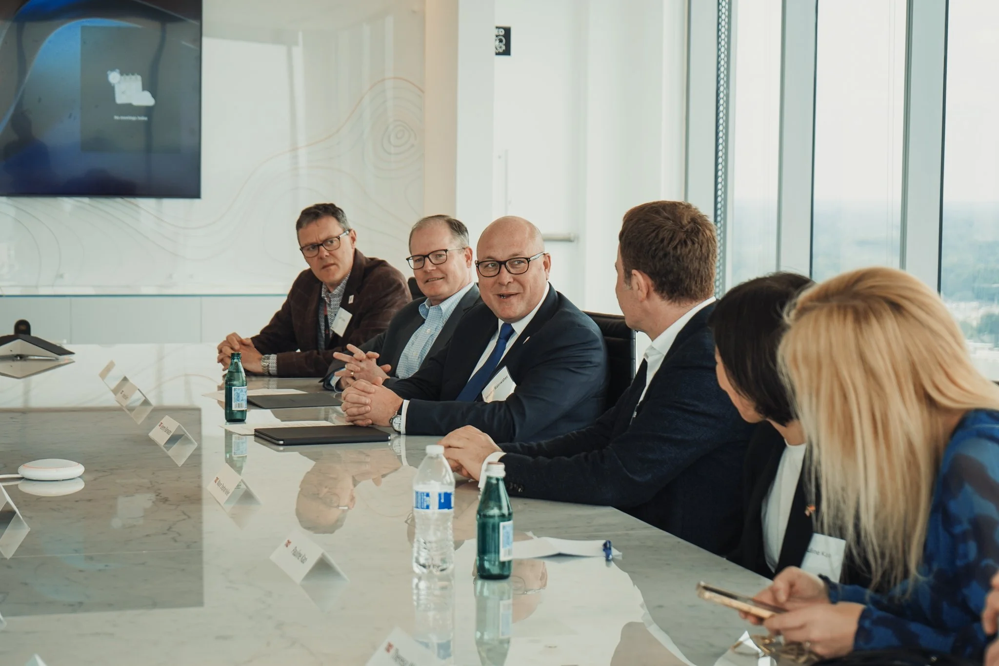A group of business professionals sitting at a conference table in a modern office, engaged in discussion, with water bottles and notepads in front of them.