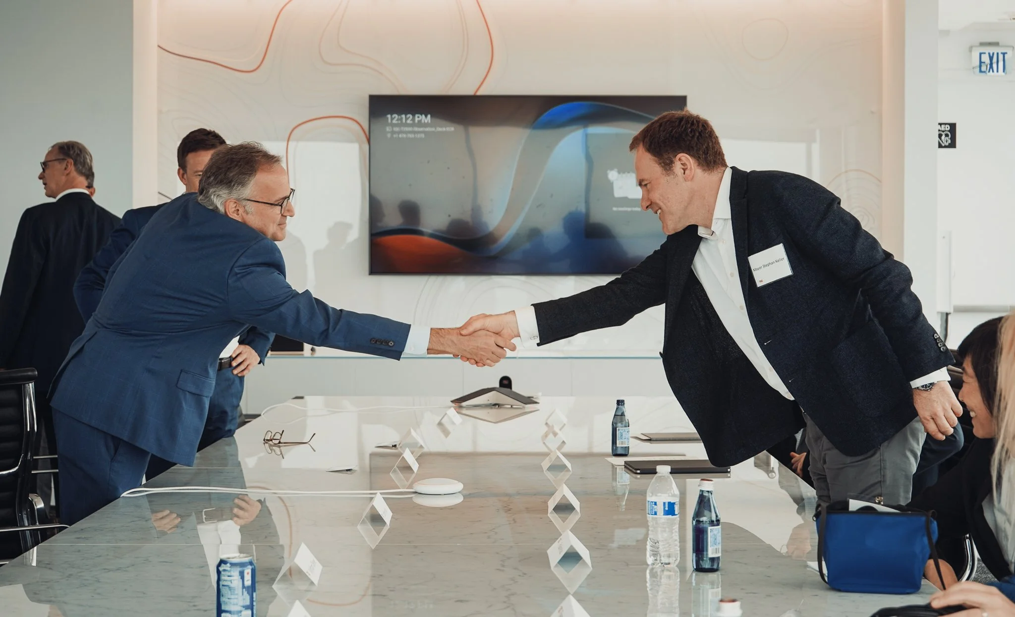 Two men are shaking hands across a conference table in a business meeting room, with several seated attendees, some standing, and a large digital display on the wall showing the time 12:12 PM.