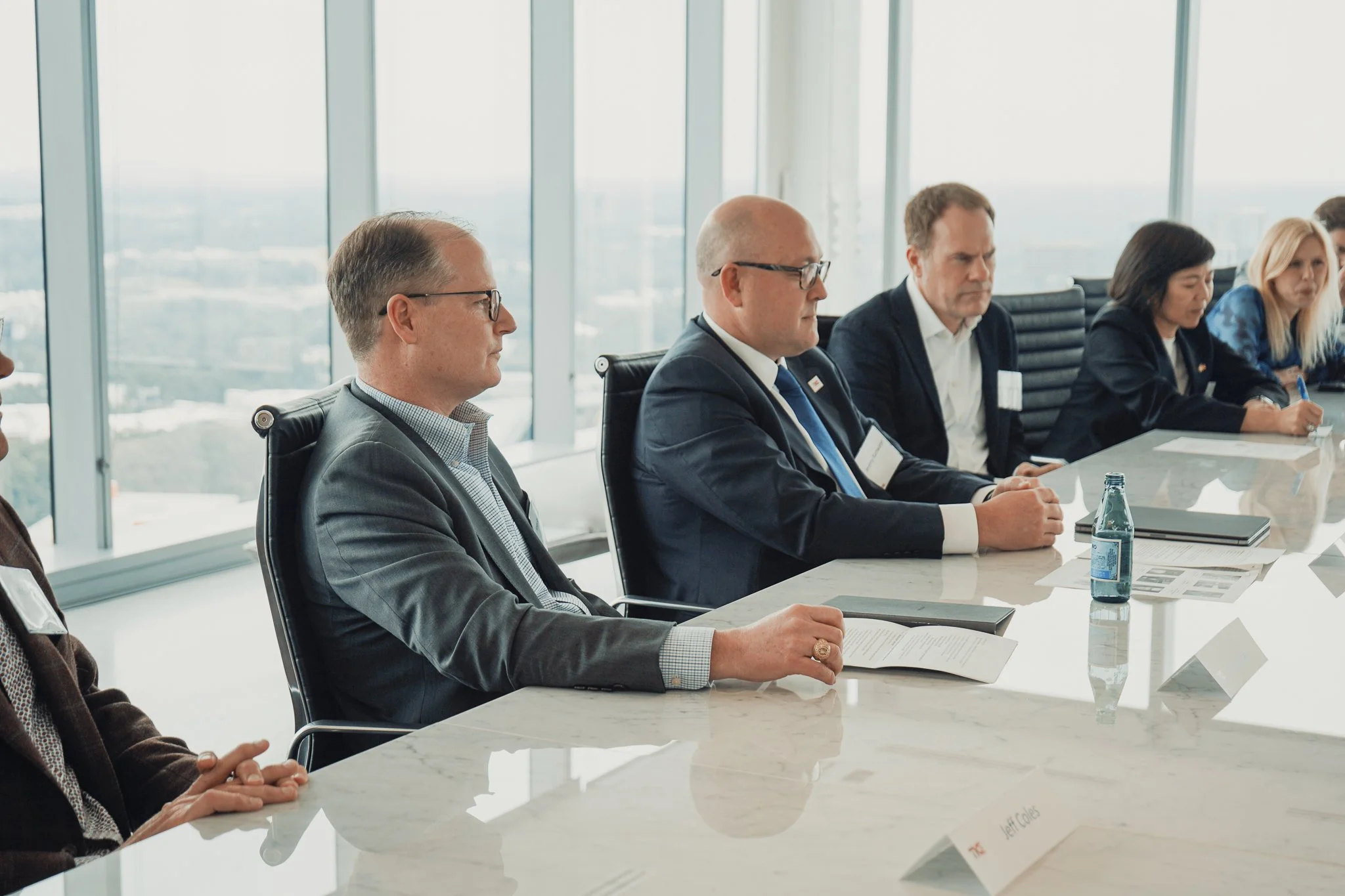 A group of businesspeople sitting at a white conference table in a modern office with large windows showing a cityscape. They are engaged in a discussion, with papers, a laptop, and bottled water on the table.