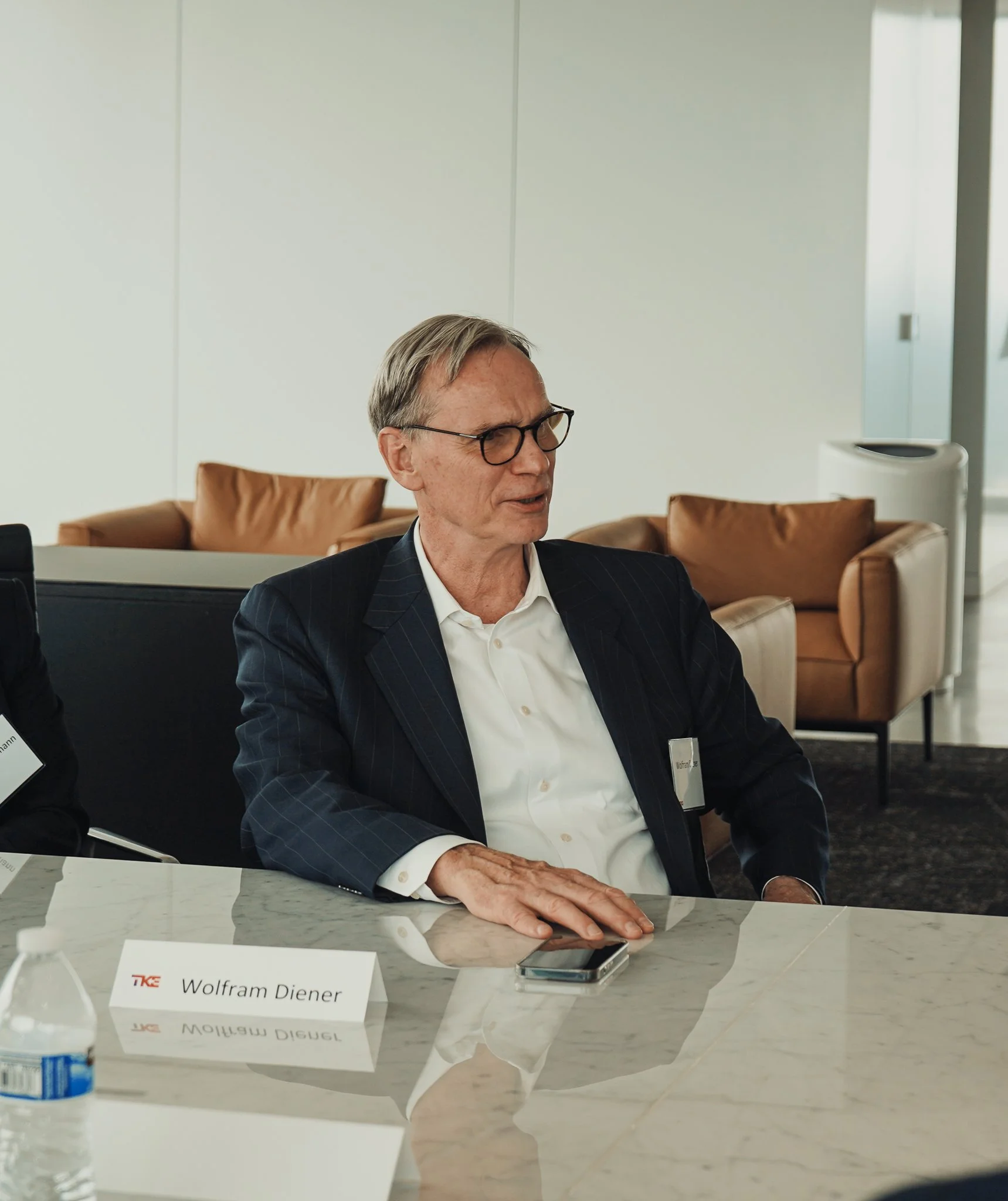 A man with glasses, wearing a dark suit and white shirt, sitting at a conference table with a nameplate that reads 'Wolfram Diener' and a cellphone, in a modern office or meeting room with beige chairs and white walls.