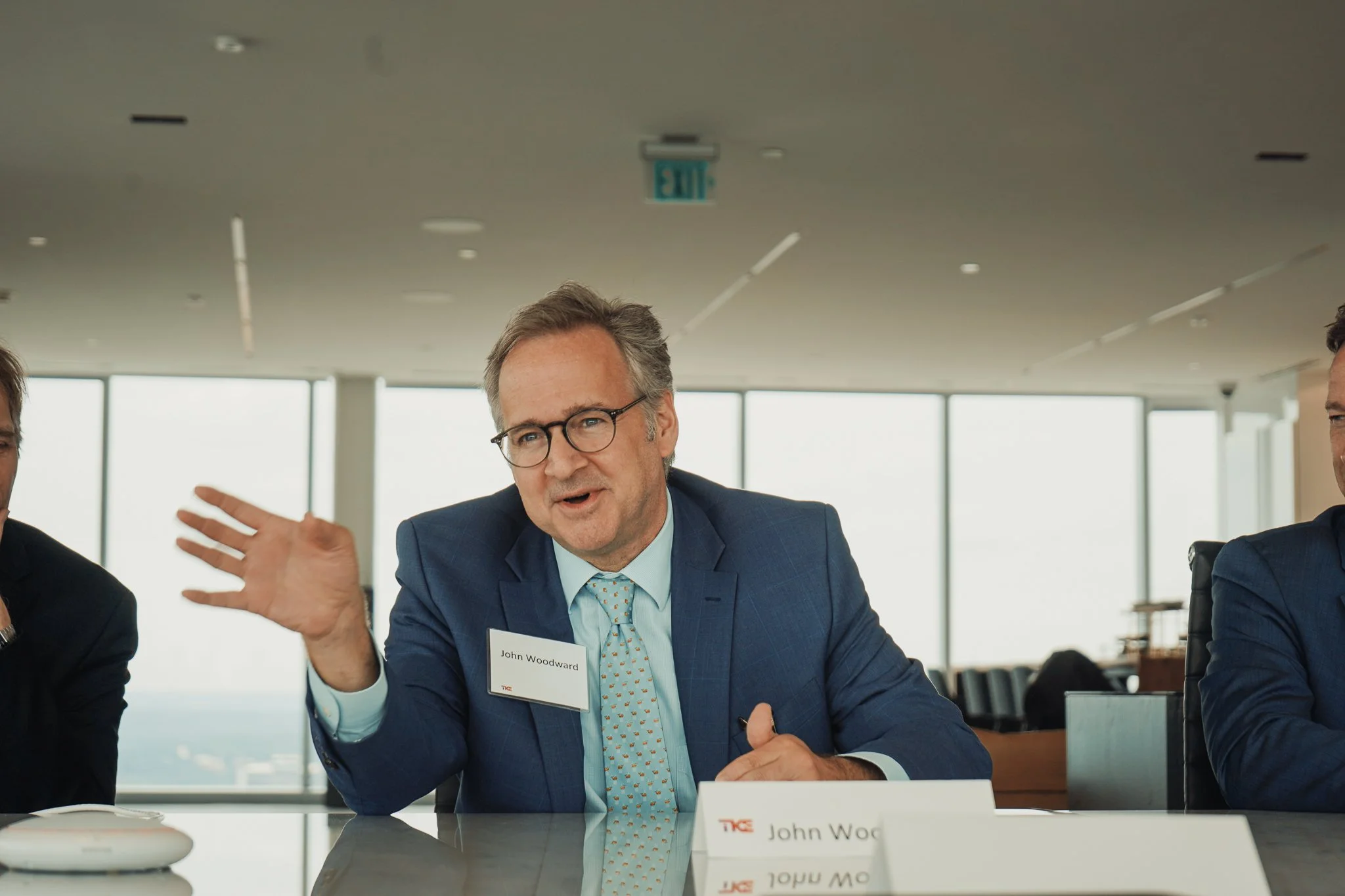 A middle-aged man with glasses, wearing a blue suit and a patterned tie, is speaking and gesturing with his hand during a business meeting in a conference room. The man has a name tag that reads 'John Woodward'.