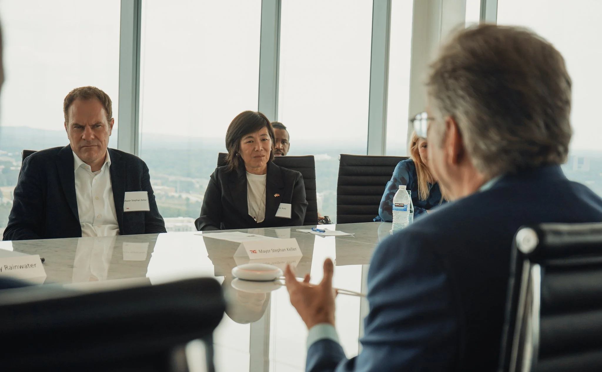 Group of business professionals in a meeting room with tall windows and city views, sitting around a table with name cards, water bottles, and papers.