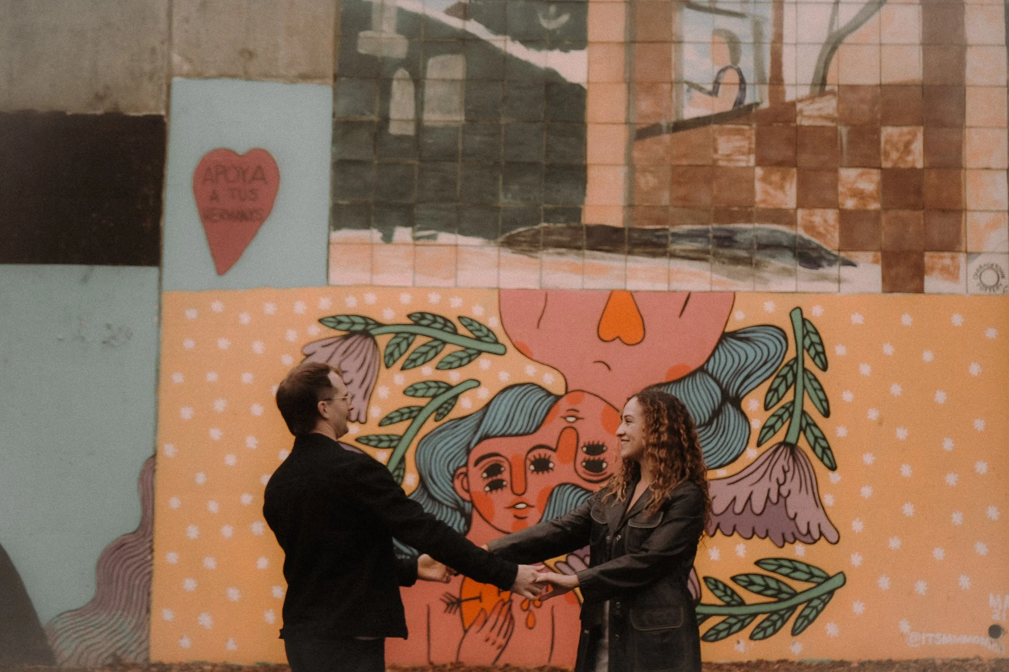 Two people, a man and a woman, are holding hands and facing each other in front of a colorful mural. The mural features artwork of a woman's face with long flowing hair, surrounded by leaves and abstract shapes.