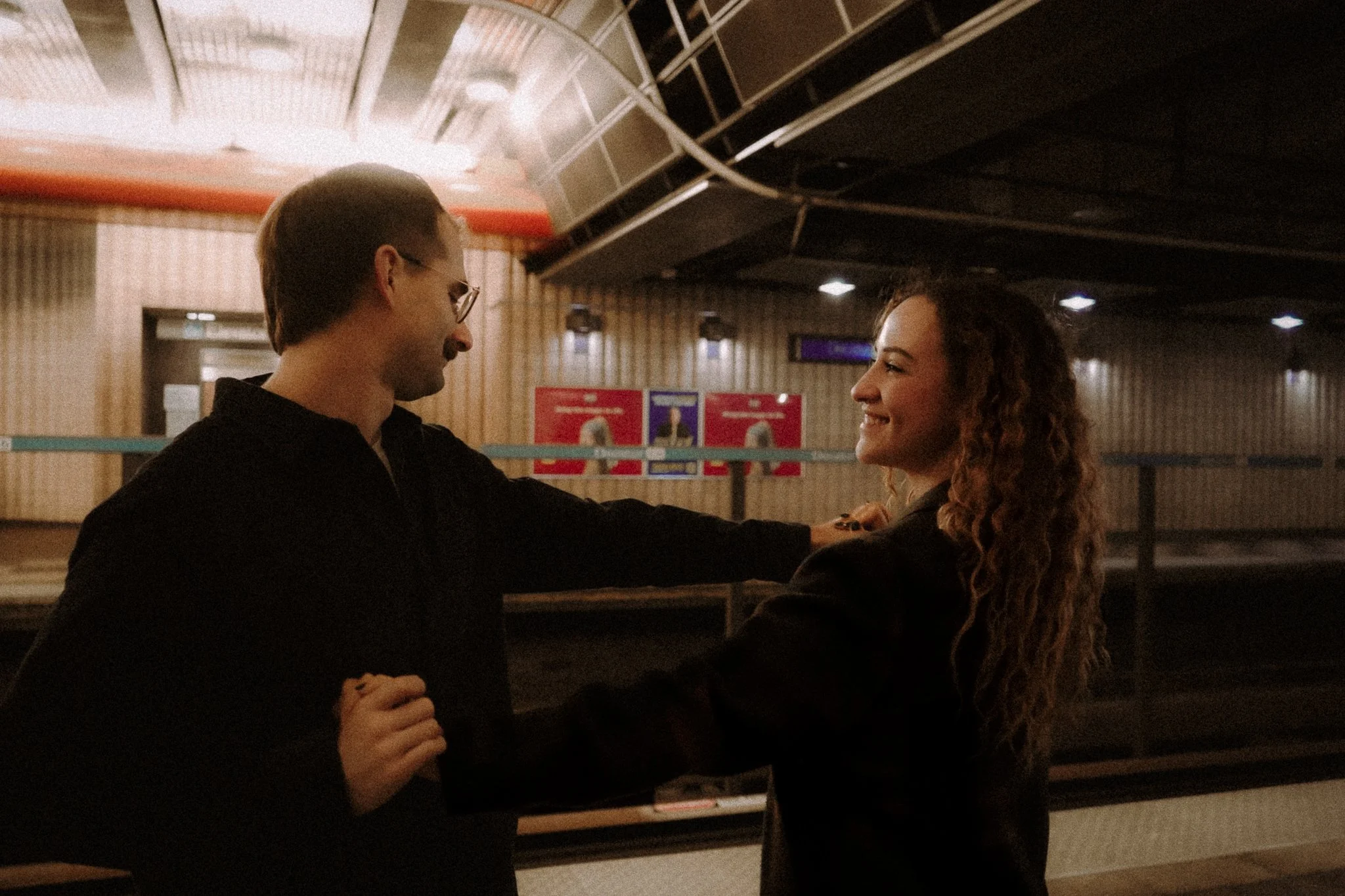Two people, a man with glasses and a woman with curly hair, are smiling and looking at each other in an indoor setting with a curved railing and posters on the wall behind them.