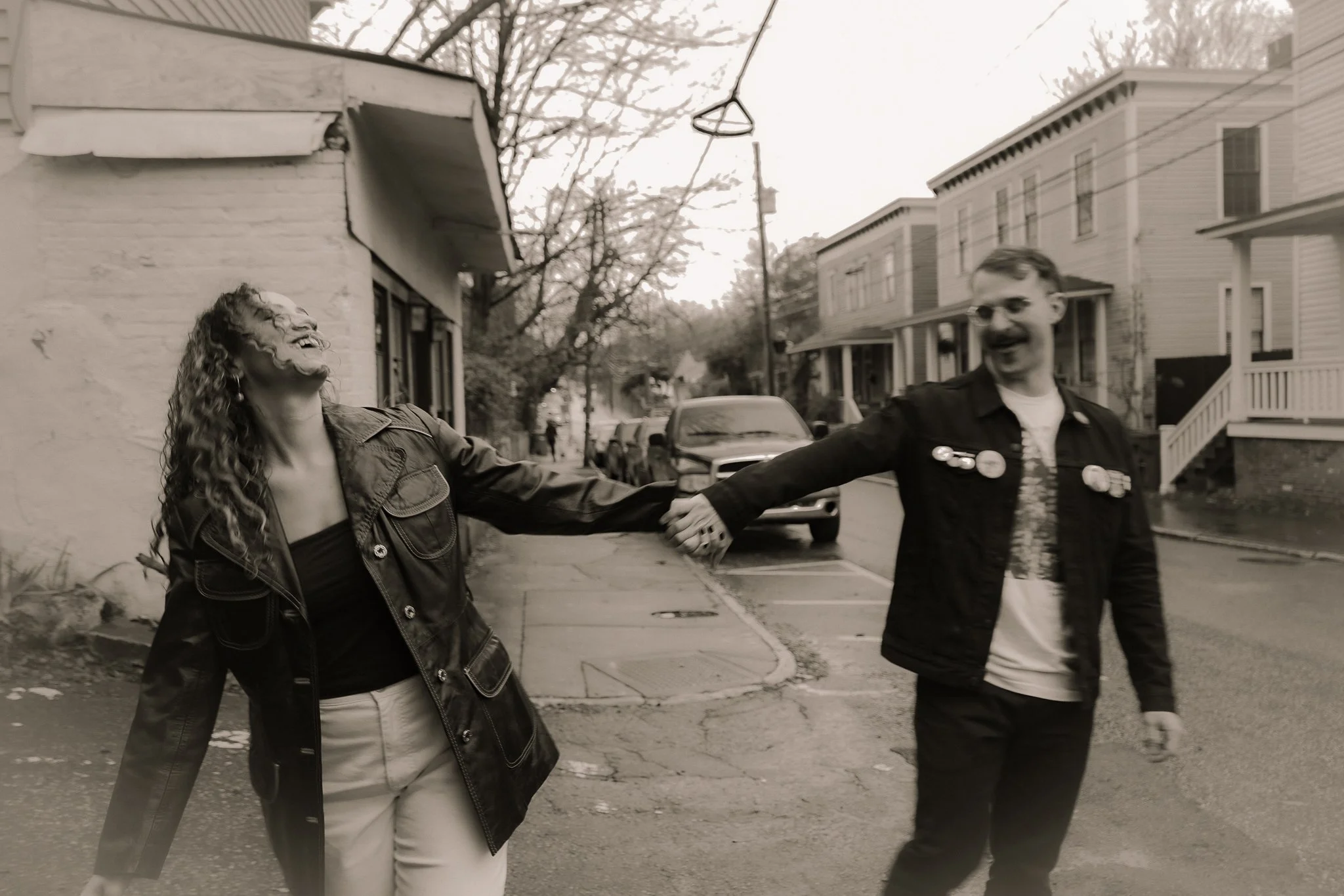 Two people smiling and holding hands outside on a city street, with vintage-style houses and parked cars in the background, black and white photo.