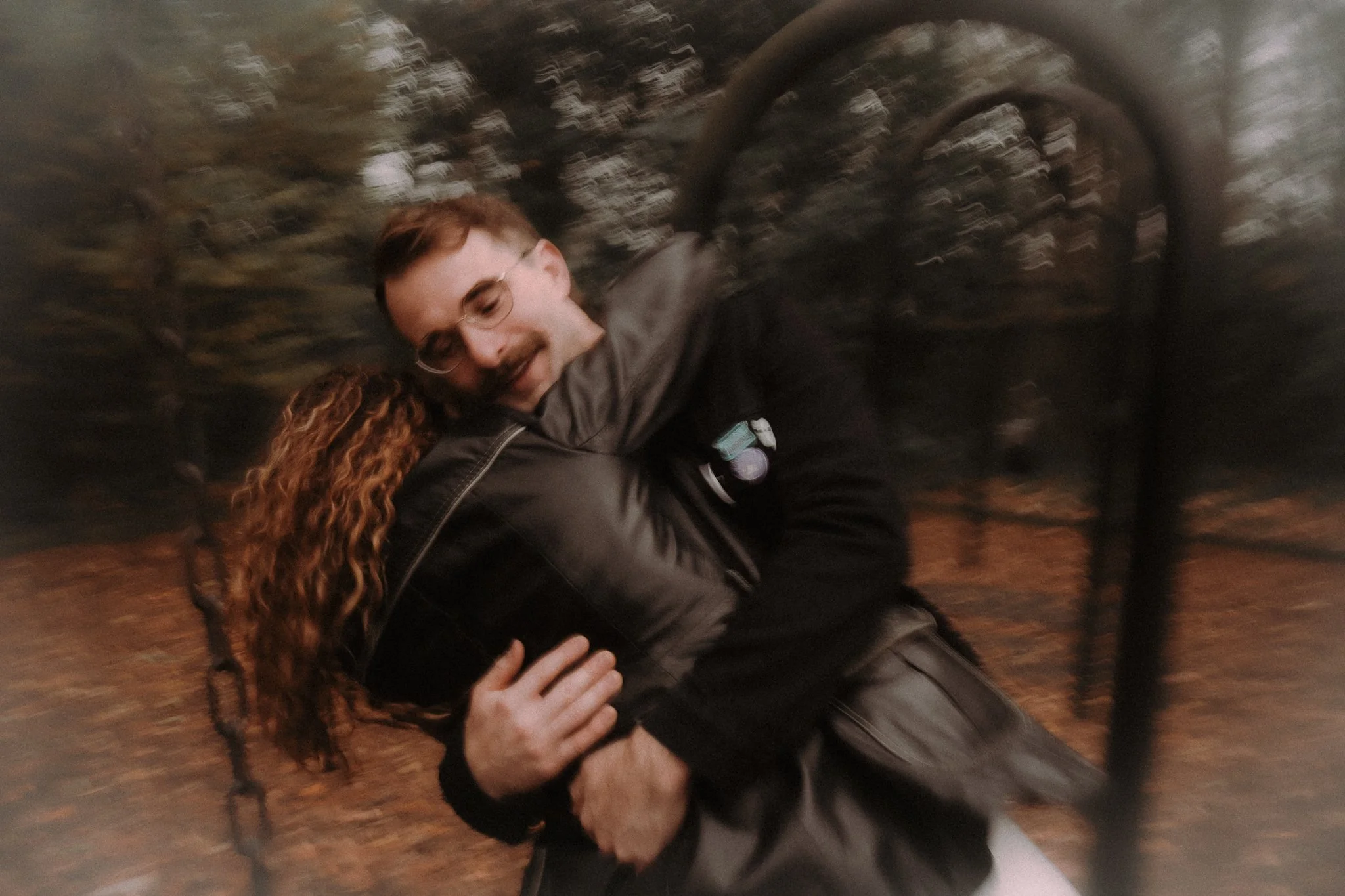 Two people hugging, one with curly red hair and a gray hoodie, and the other with spiky hair and a black jacket, in a blurry outdoor setting.