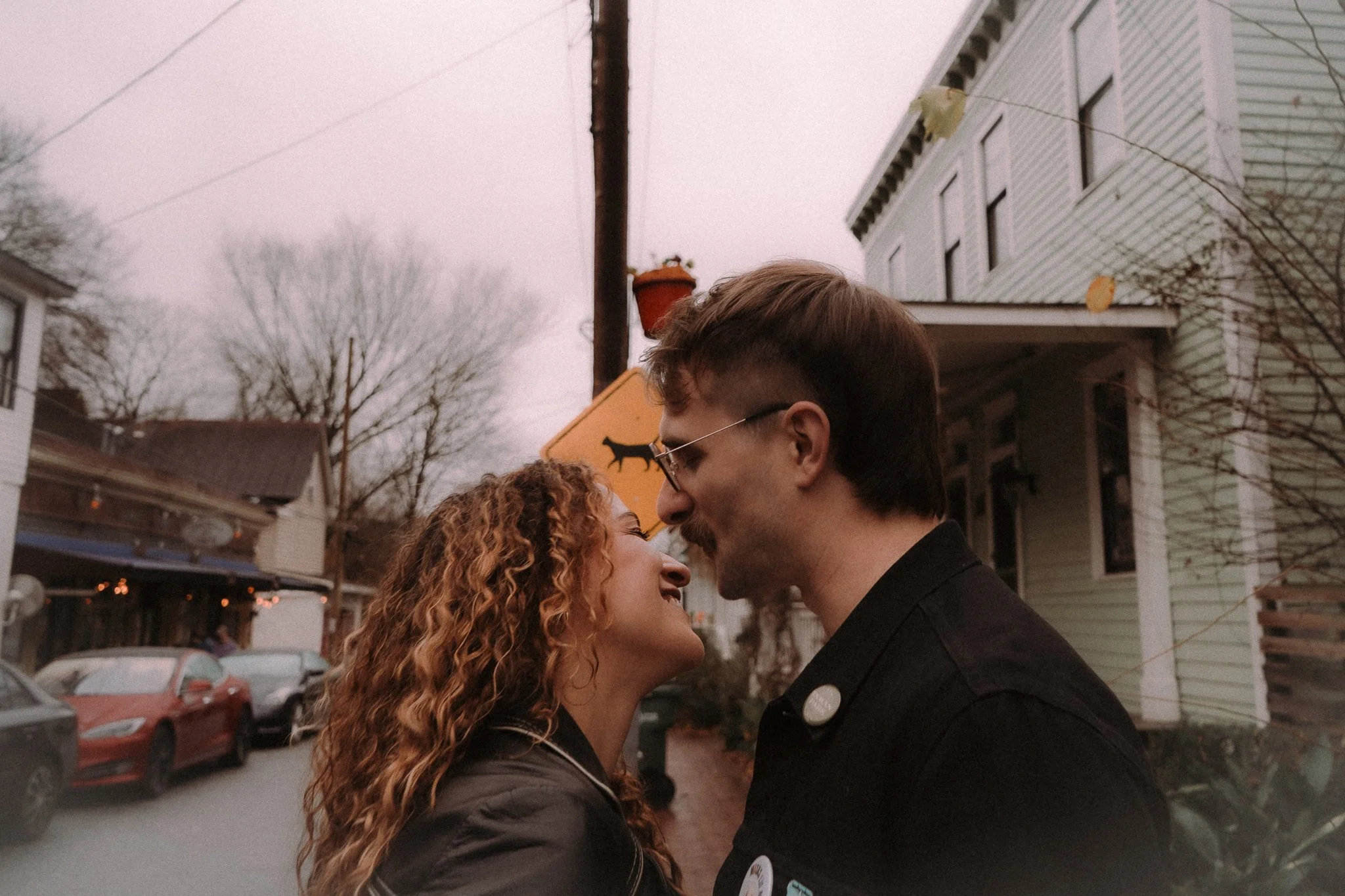 A couple standing close and facing each other outdoors in a neighborhood, with trees, cars, and houses in the background, as clouds gather overhead.