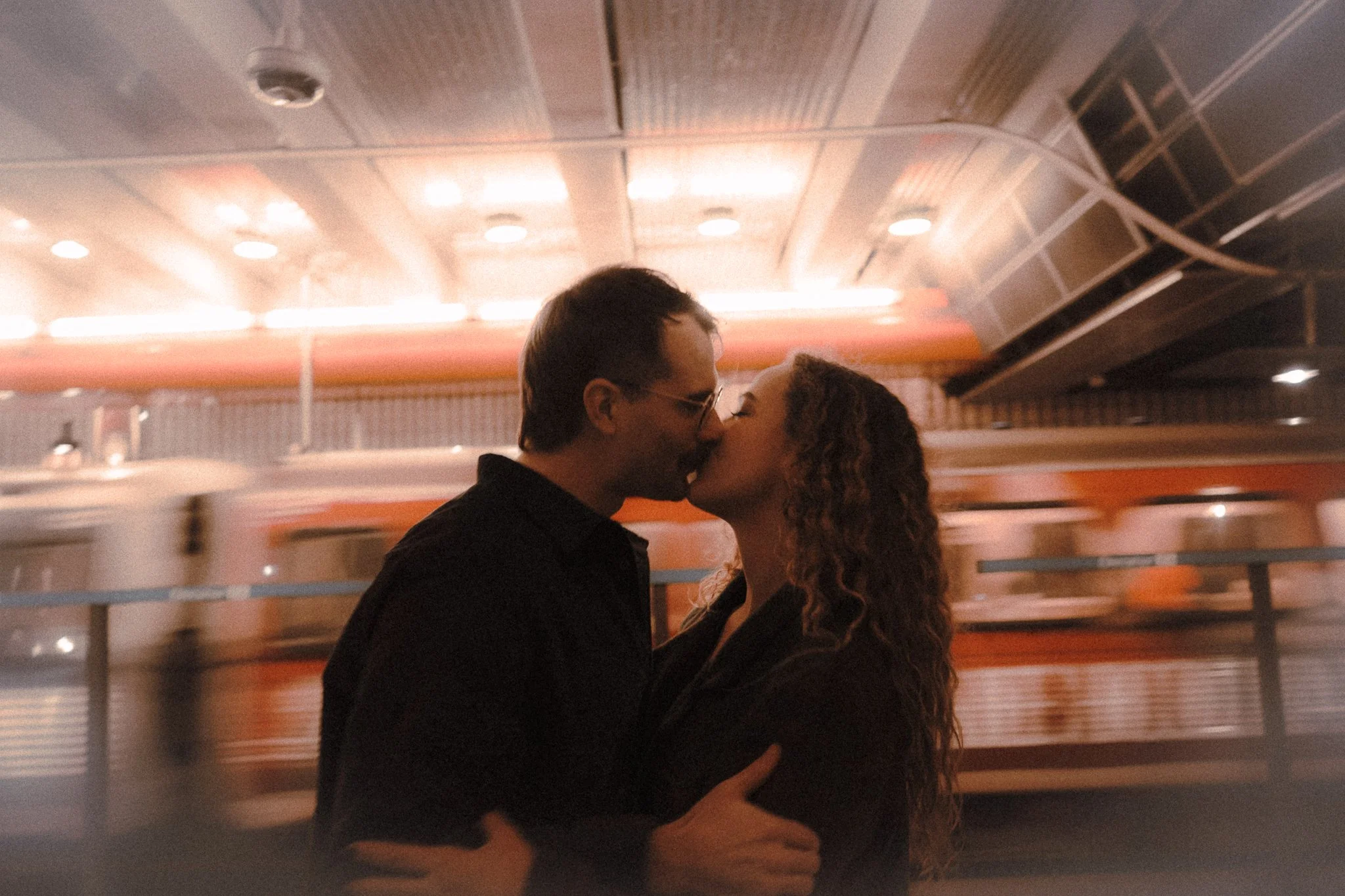 A couple kissing in a dimly lit setting with blurred background of a ceiling with lights and a moving bus.