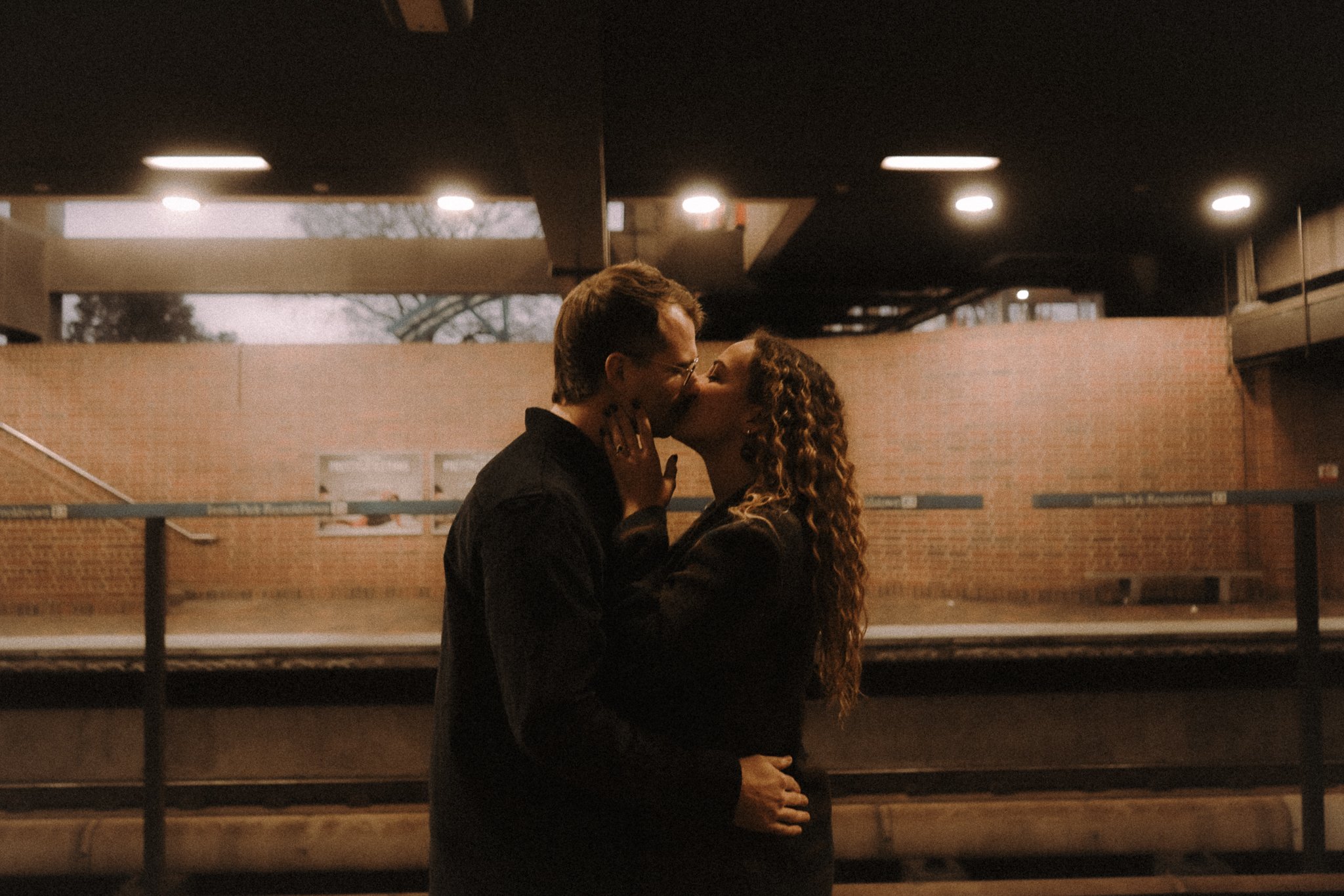 A couple sharing a kiss in an underground parking garage at sunset, with a brick building and trees in the background.