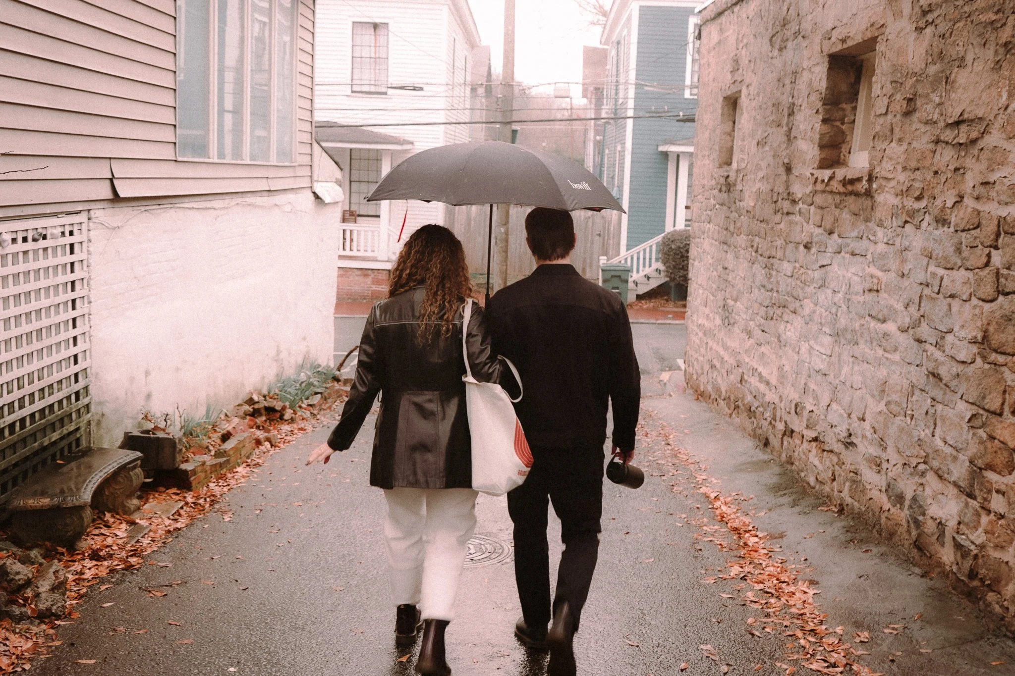 Two people, a woman and a man, walking in a narrow alleyway holding a black umbrella during rainy weather. The woman has curly hair and is wearing a leather jacket, and the man is holding a camera in his hand.