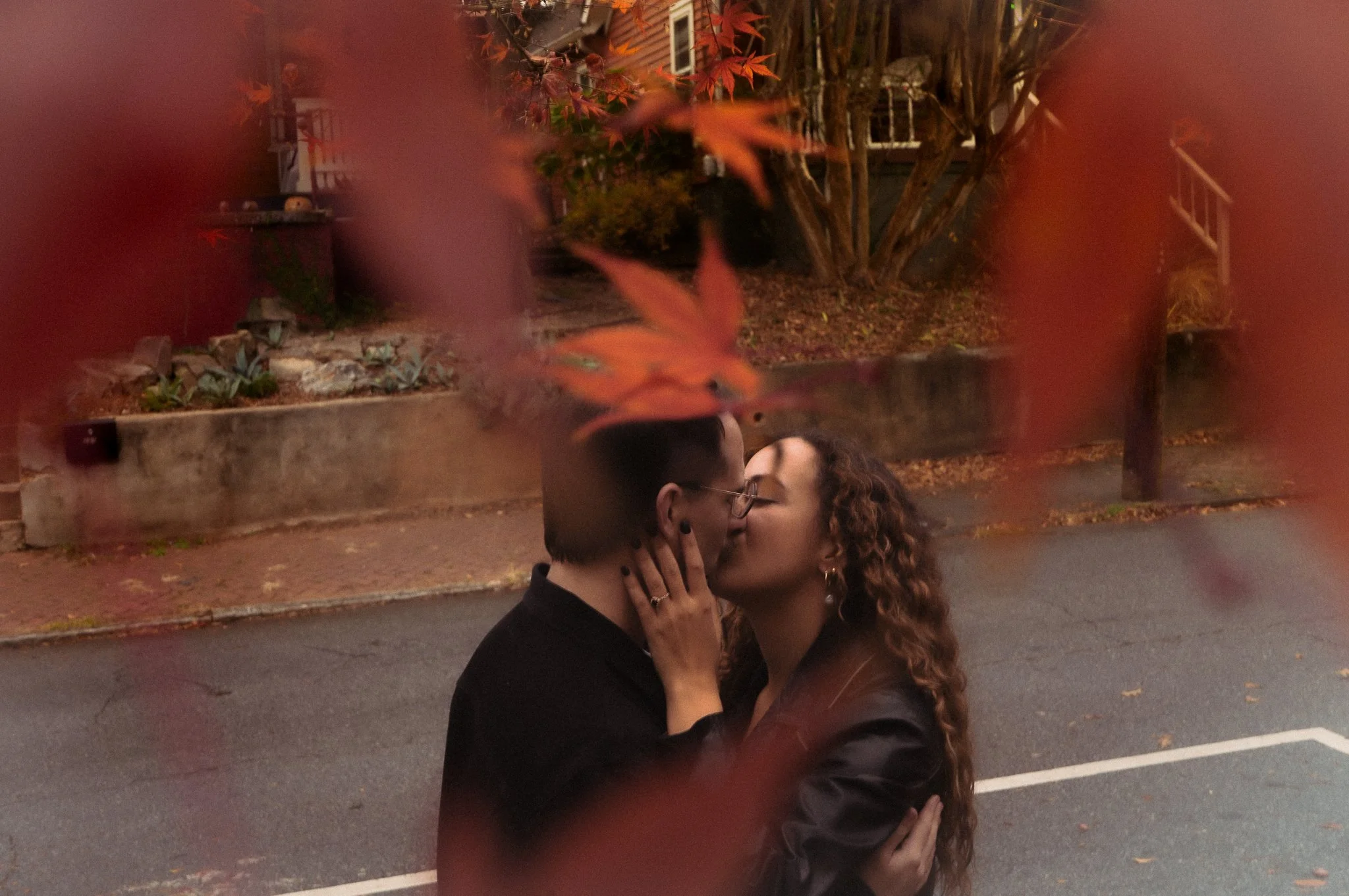 A couple is kissing on a street, captured through partly blurred red and orange autumn leaves in the foreground. The man has short dark hair and glasses and is wearing a black coat, and the woman has curly hair and is also wearing a dark coat.