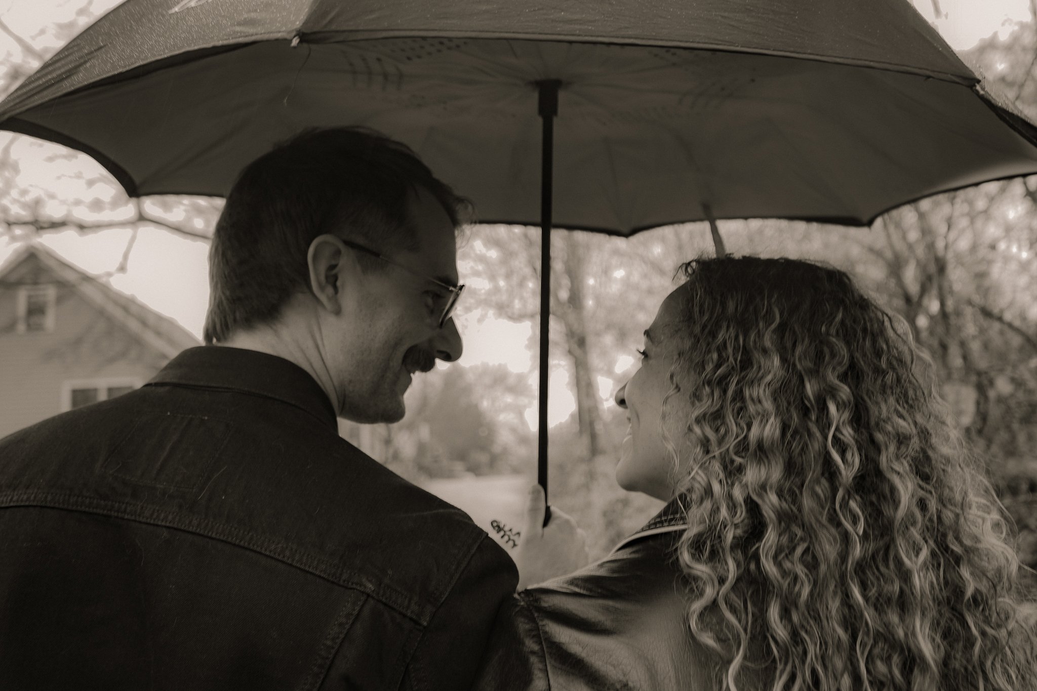 A black and white photo of a man and a woman under an umbrella, close and facing each other, outdoors during what appears to be a rainy day.