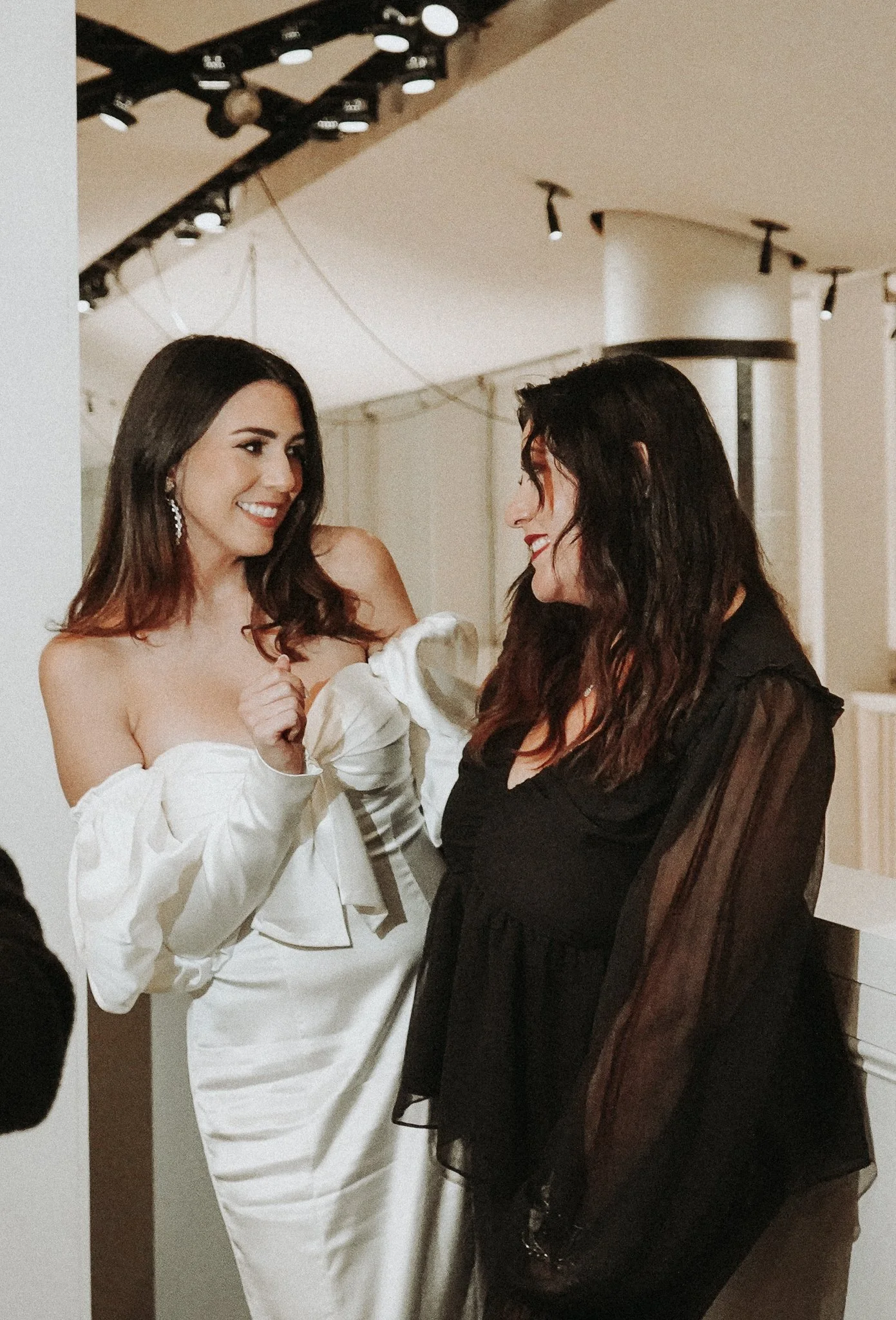 Two women are smiling and talking to each other indoors. The woman on the left has long dark hair, accessorized with earrings, and is wearing an off-the-shoulder white dress with puffy sleeves. The woman on the right has wavy dark hair and is dressed in black, with sheer long sleeves.