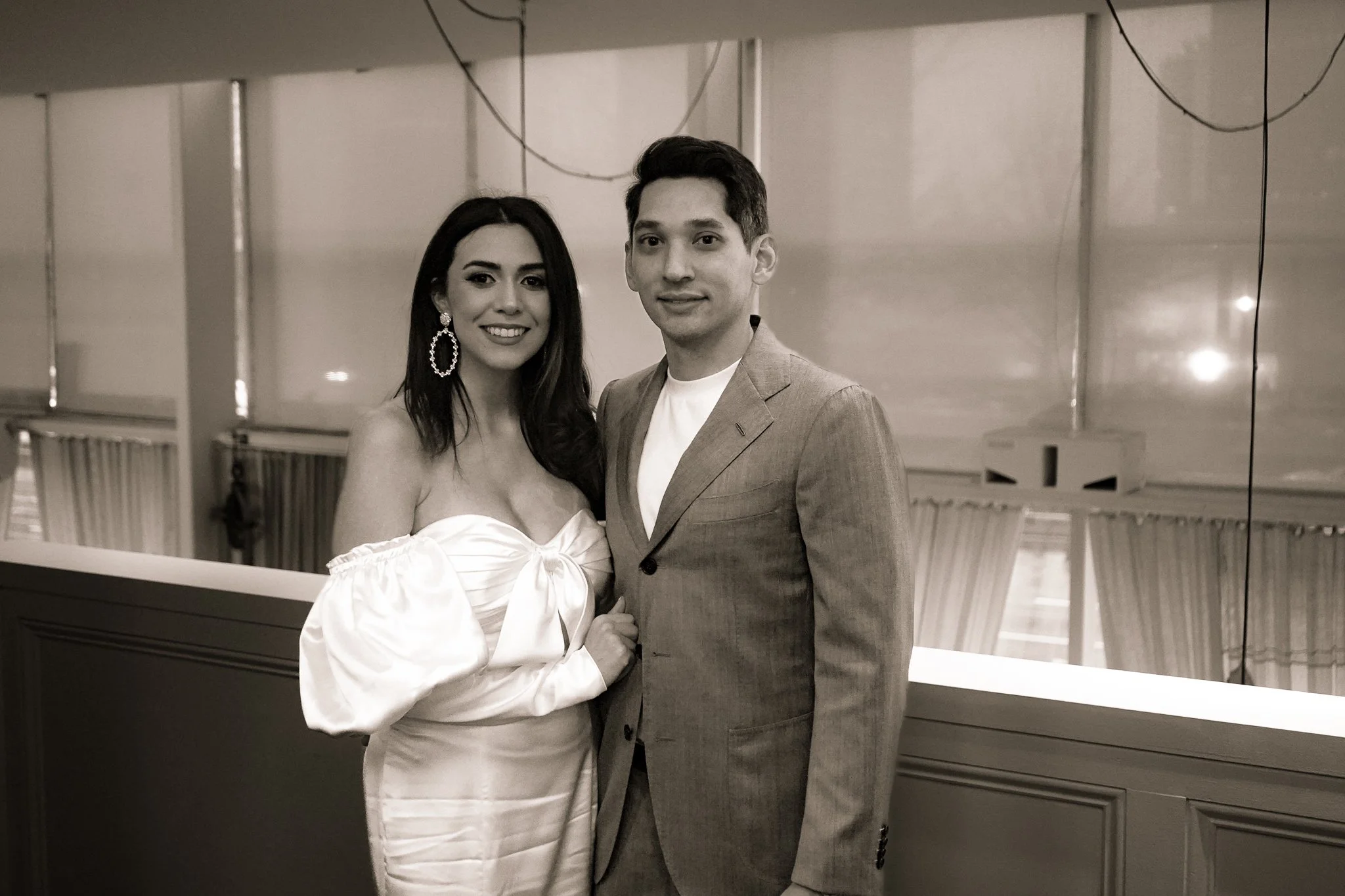 A black and white photo of a man and woman standing together indoors, smiling at the camera. The woman has long dark hair, is wearing large earrings, and a puffed sleeve dress. The man has short dark hair, is dressed in a light-colored suit jacket and white shirt. The background shows a mirrored wall with wires hanging and a piano.