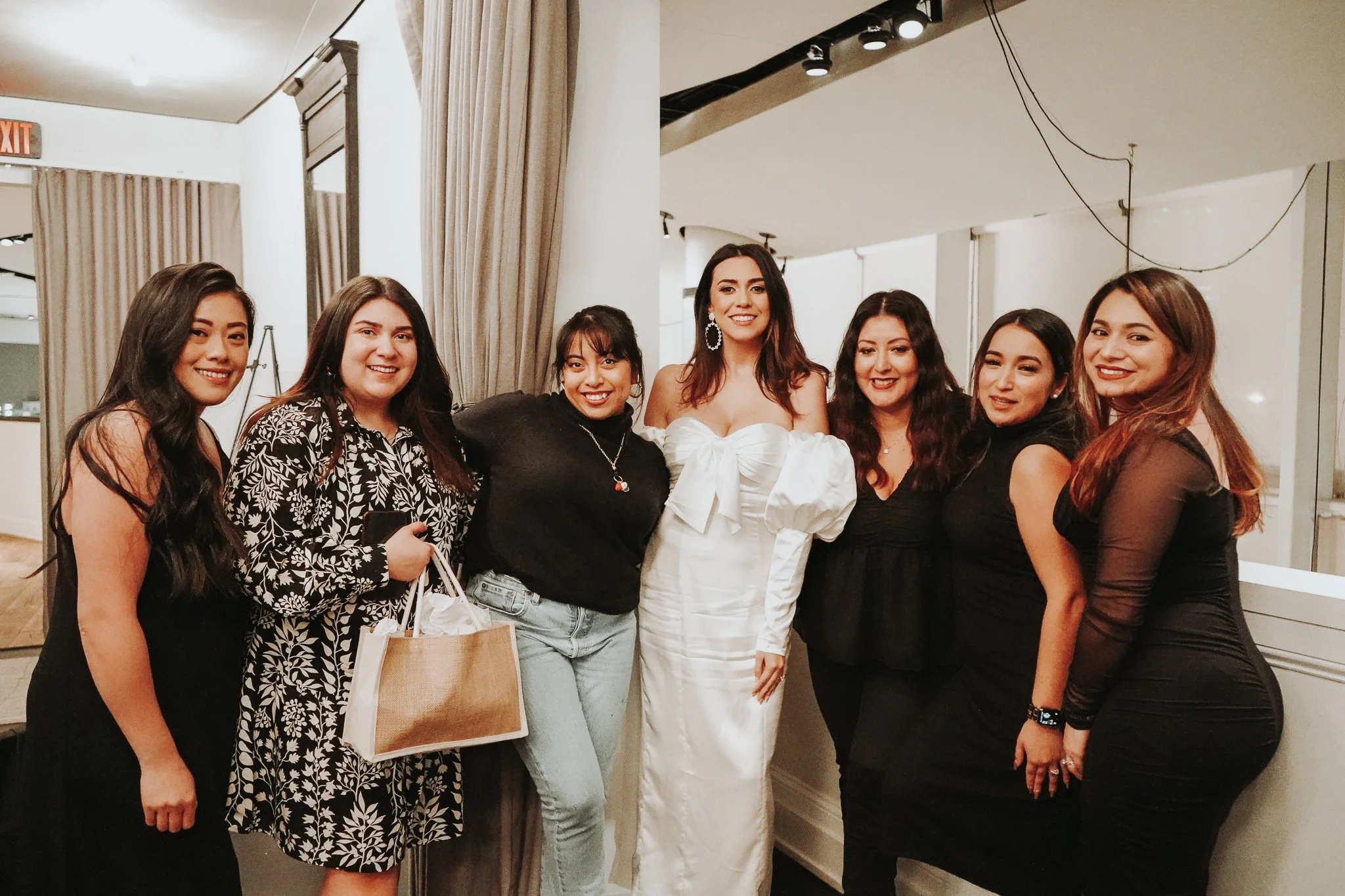 Group of seven women standing together indoors, smiling, with one woman wearing a white dress in the center.