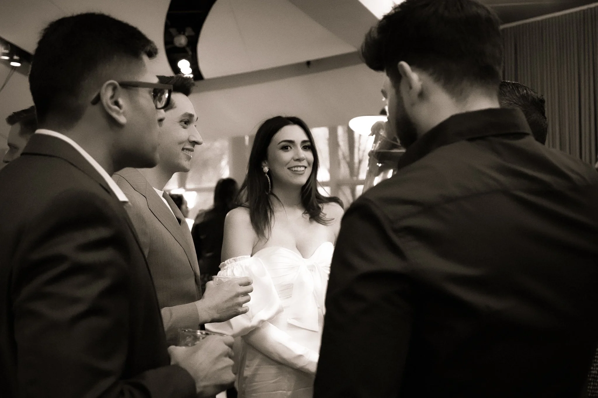Group of people at a social event, engaged in conversation, with a woman in a white off-shoulder dress smiling in the center.