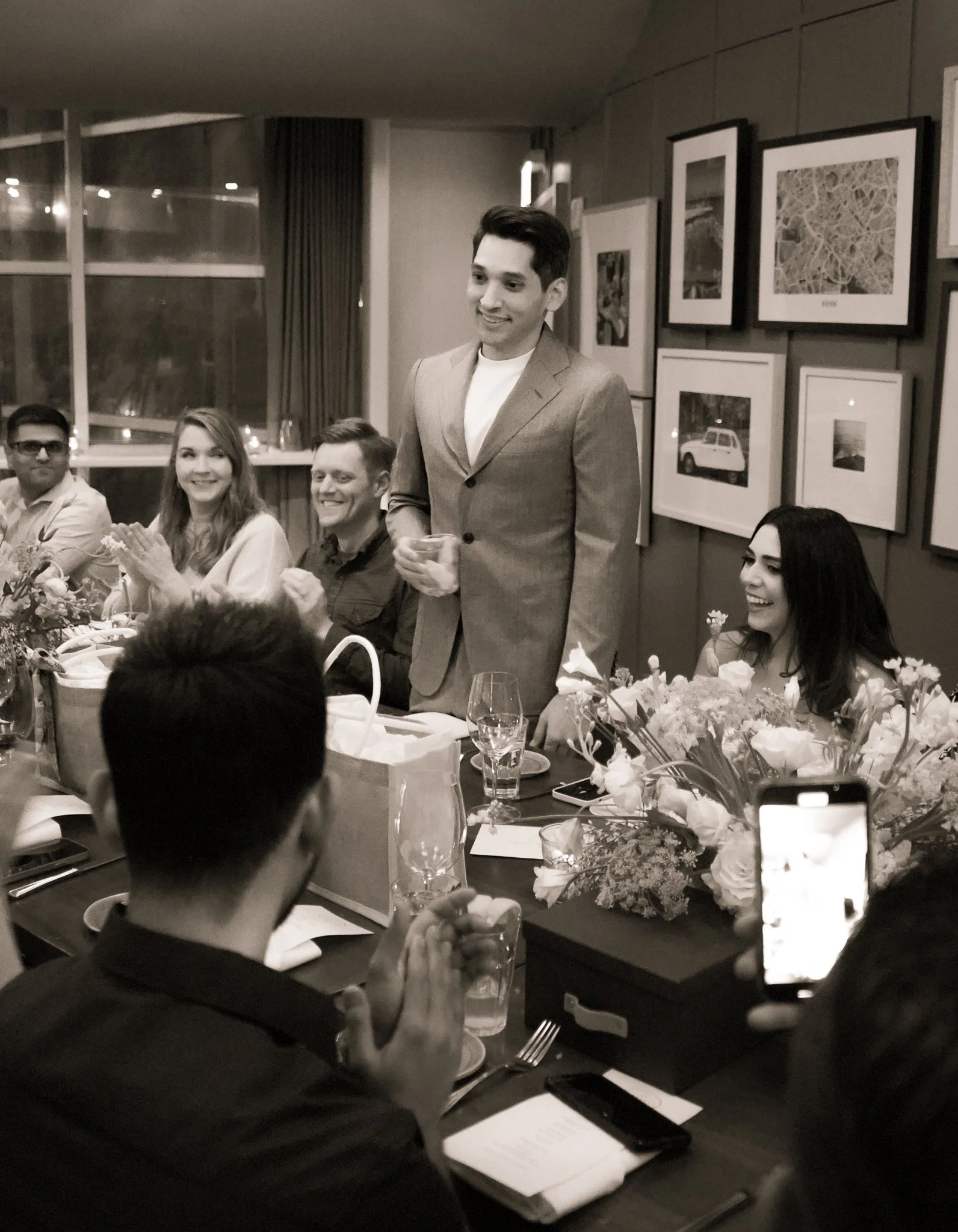 A group of people attending a formal dinner, with some clapping and smiling, as a man in a suit is giving a speech or toast at the head of the table. The table is decorated with flowers, and there are framed photos on the wall in the background.