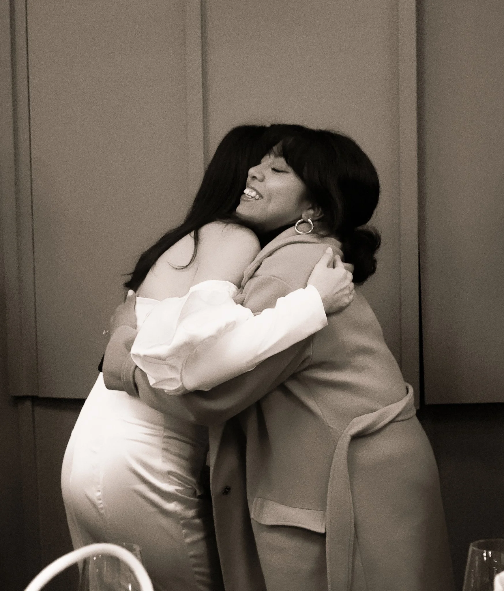 Two women hugging in a close embrace, smiling with eyes closed, in a room with plain walls, one wearing earrings and a blazer, the other with long hair and white shirt.
