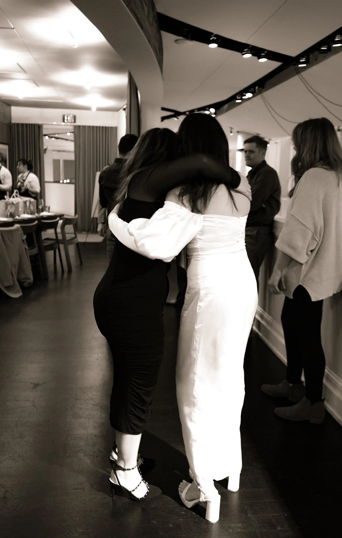Two women hug at a social gathering, one dressed in black, the other in white with high-heeled shoes, in a room with other standing and sitting people.