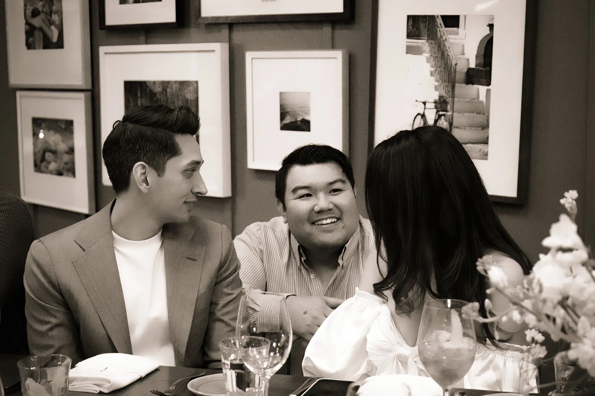 Three people sitting at a table in a restaurant, engaged in conversation. The wall behind them is decorated with black-and-white framed photos, including landscapes and architectural elements. The table has glasses of water, wine, napkins, and a plat