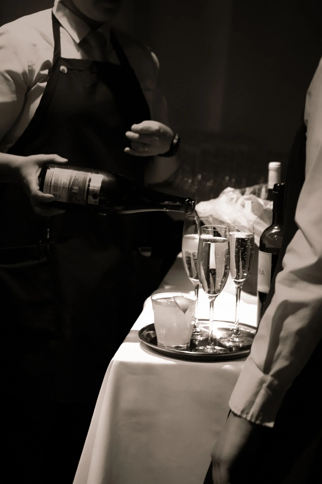 Person pouring a beverage into a glass on a table set with multiple champagne glasses, a tray, and bottles, at a formal event.