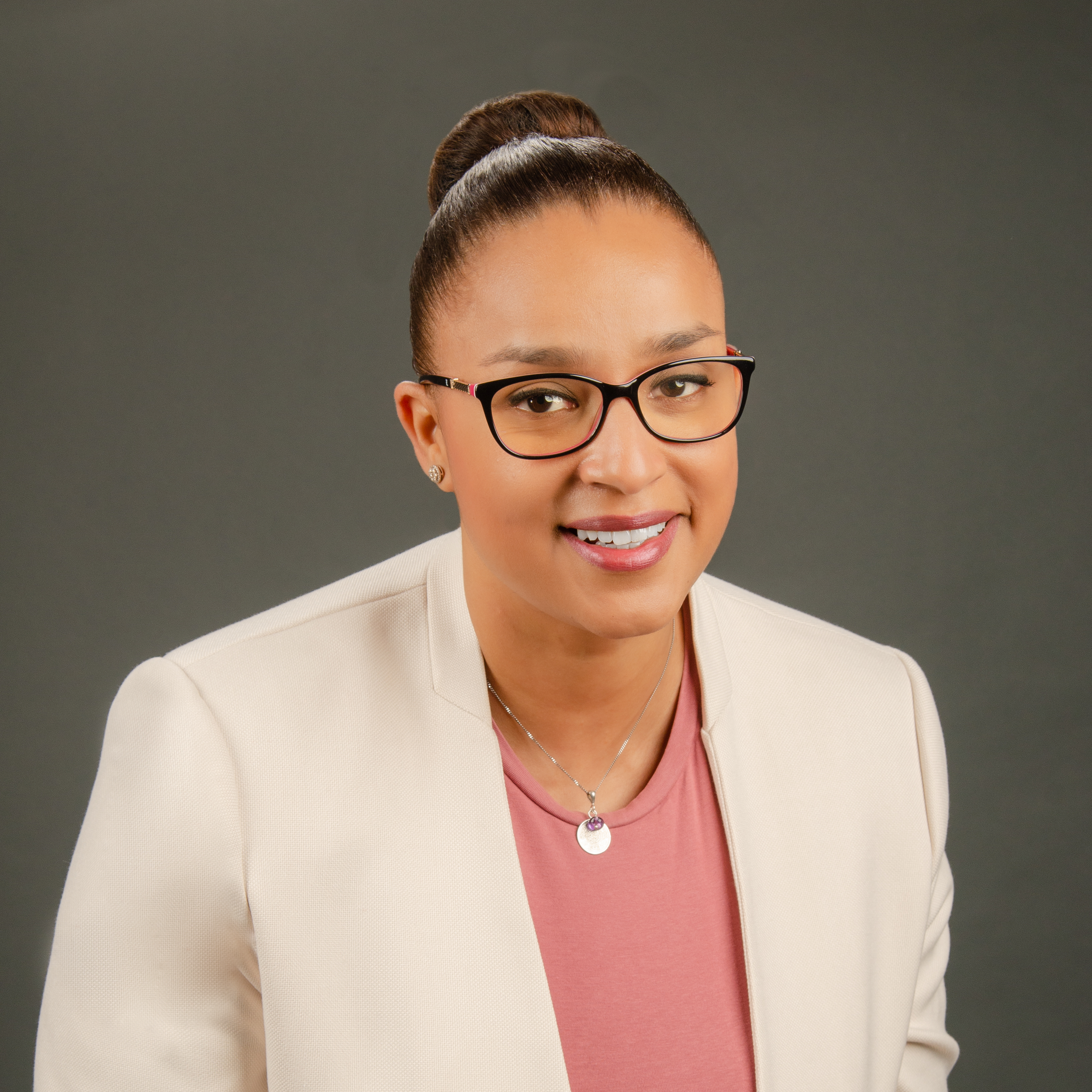 Close-up portrait of a smiling woman with glasses, wearing a beige blazer and pink top, against a gray background.