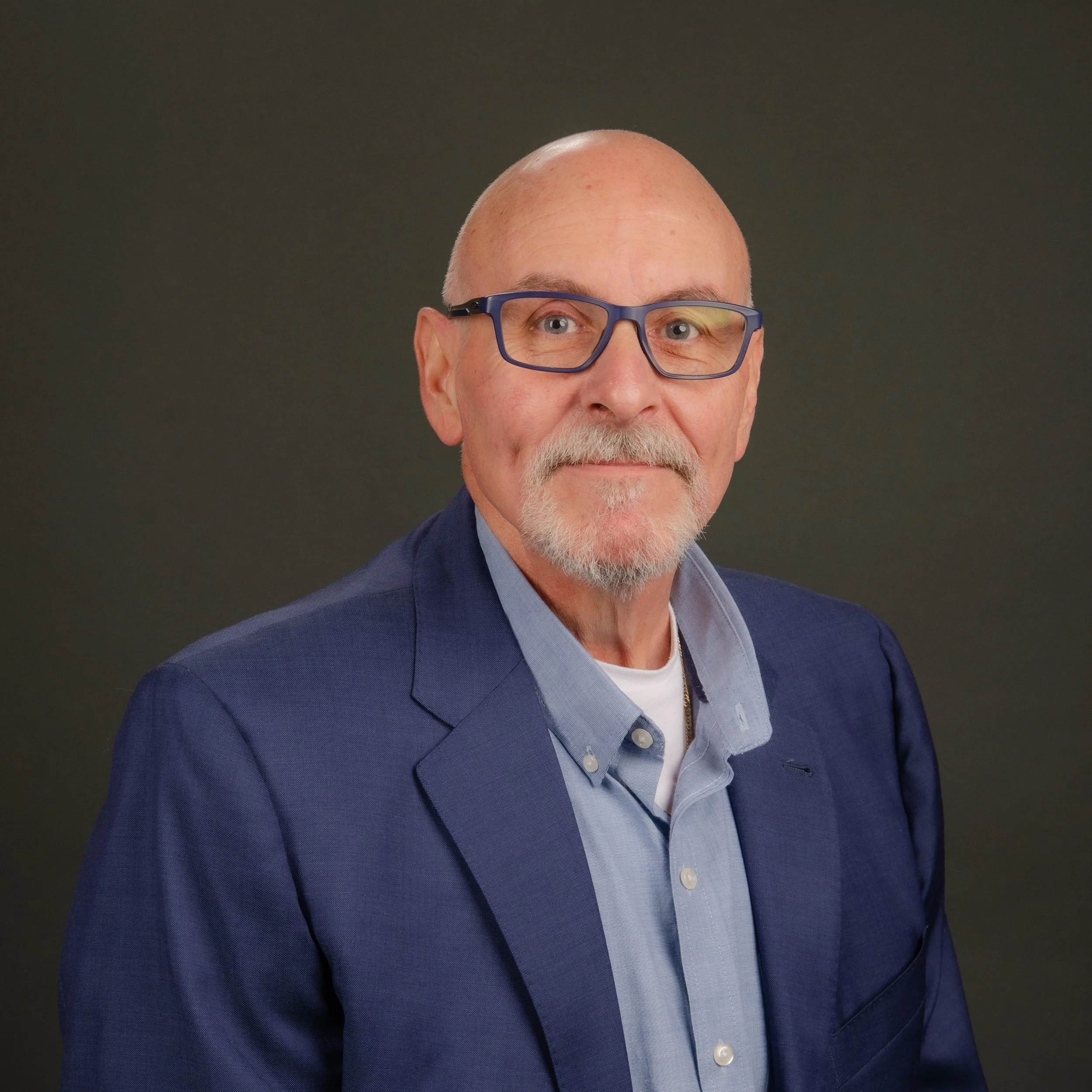 Close-up of an elderly man with a bald head and white beard wearing blue framed glasses and a blue suit against a dark background.