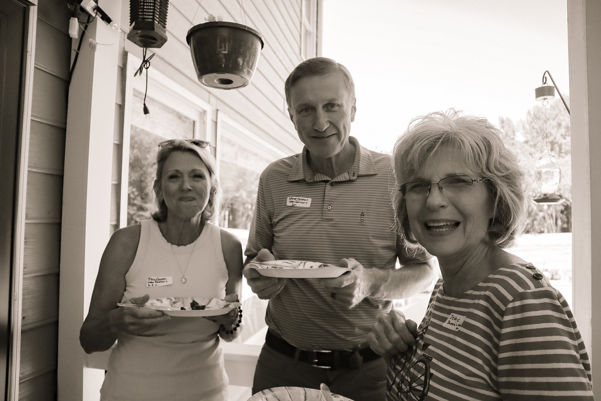 Group of four adults standing outside on a porch, smiling and holding plates of food, with a porch light and siding visible in the background, black and white photo.