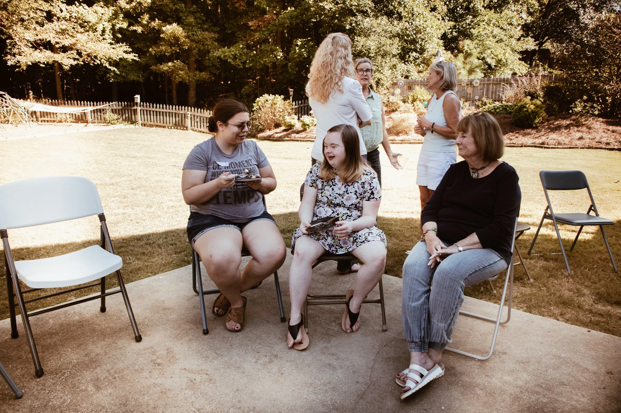 Group of women and a girl talking and sitting outdoors in a backyard garden with trees and fencing, some sitting on chairs and others standing, enjoying a sunny day.