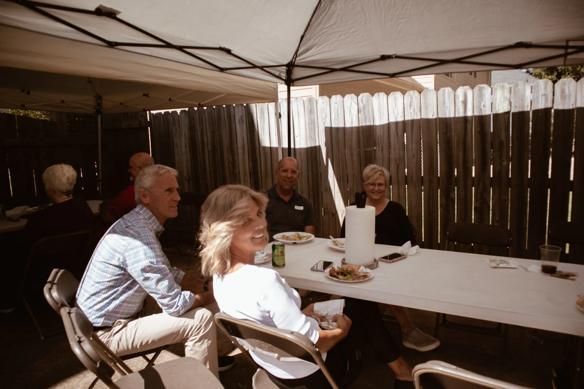 A group of people sitting at a picnic table outdoors on a sunny day, under a canopy, with food, drinks, and personal items on the table. They are smiling and engaging with each other, with a wooden fence in the background.