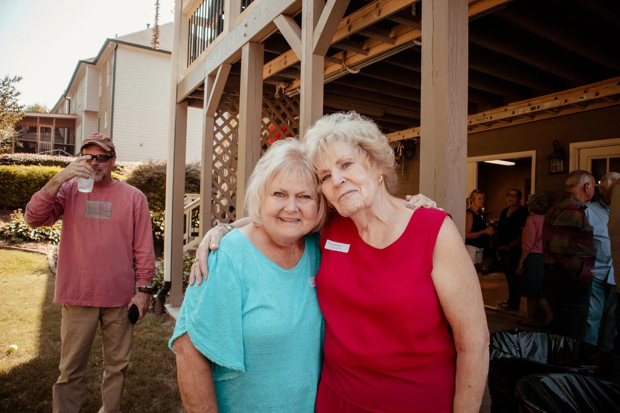 Two elderly women hugging and smiling at an outdoor gathering, with a man drinking water in the background, in a backyard setting during daytime.