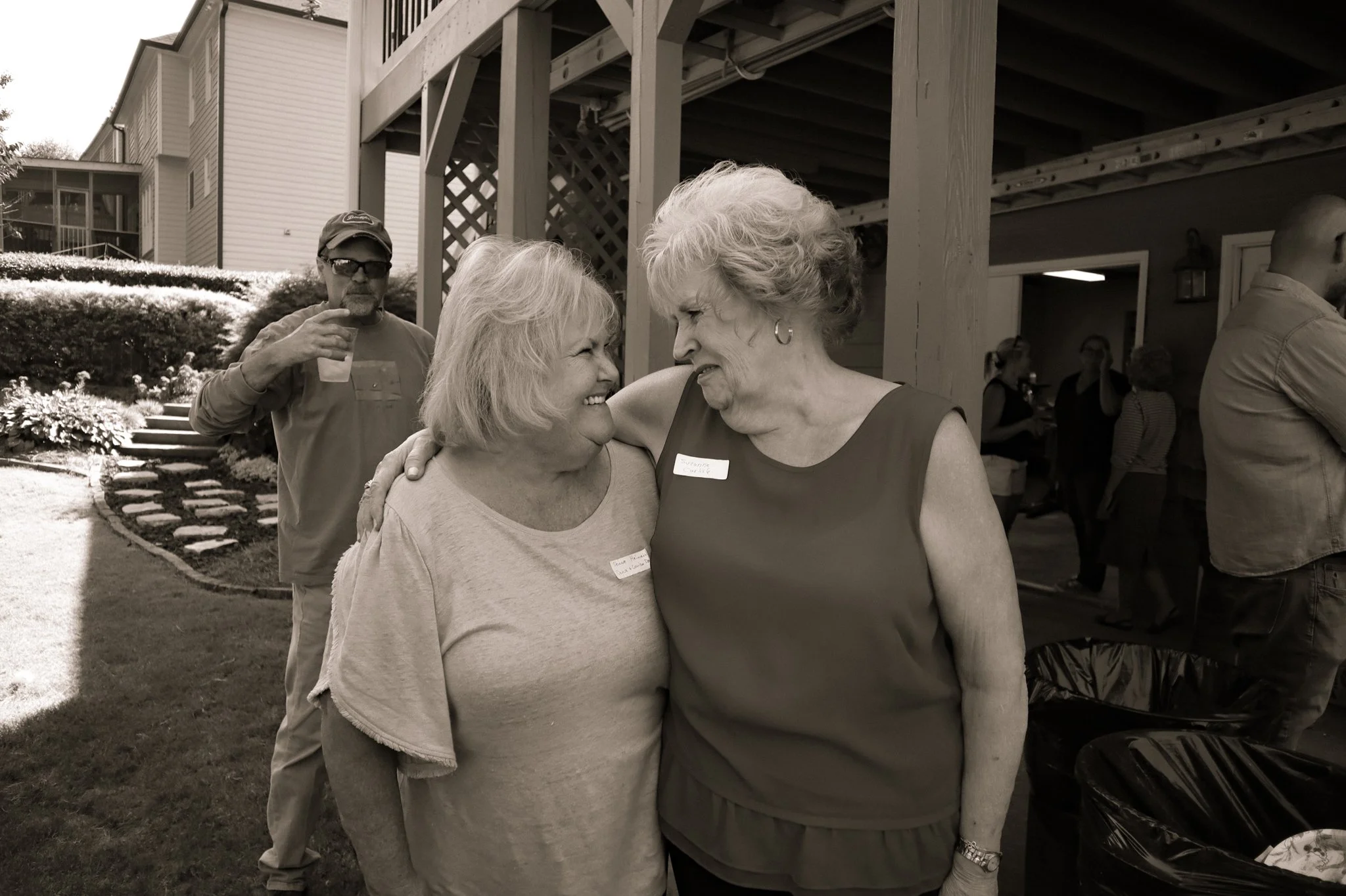 Two women are embracing and smiling at each other during an outdoor gathering, with other people in the background.