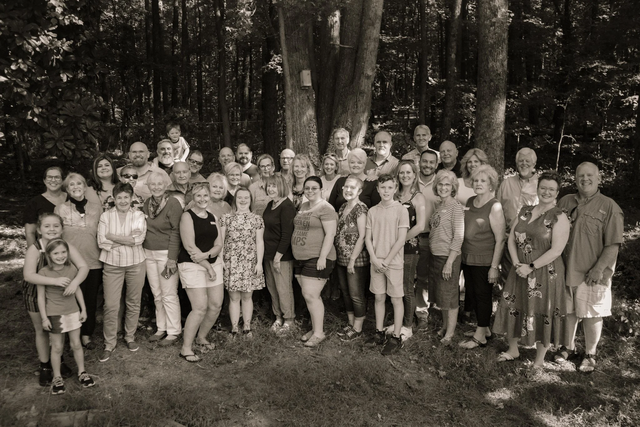 A large group of people of various ages standing outdoors in front of trees, posing for a group photo in a forested area.