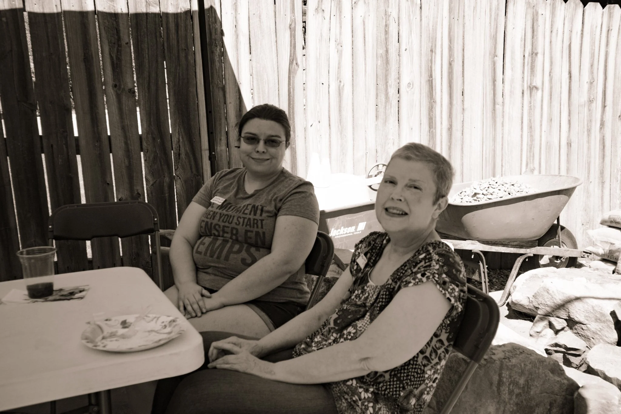 Two women sitting at a table outdoors, with a wooden fence in the background, one wearing glasses and a t-shirt, and the other wearing a patterned shirt, among scattered plates and glasses.