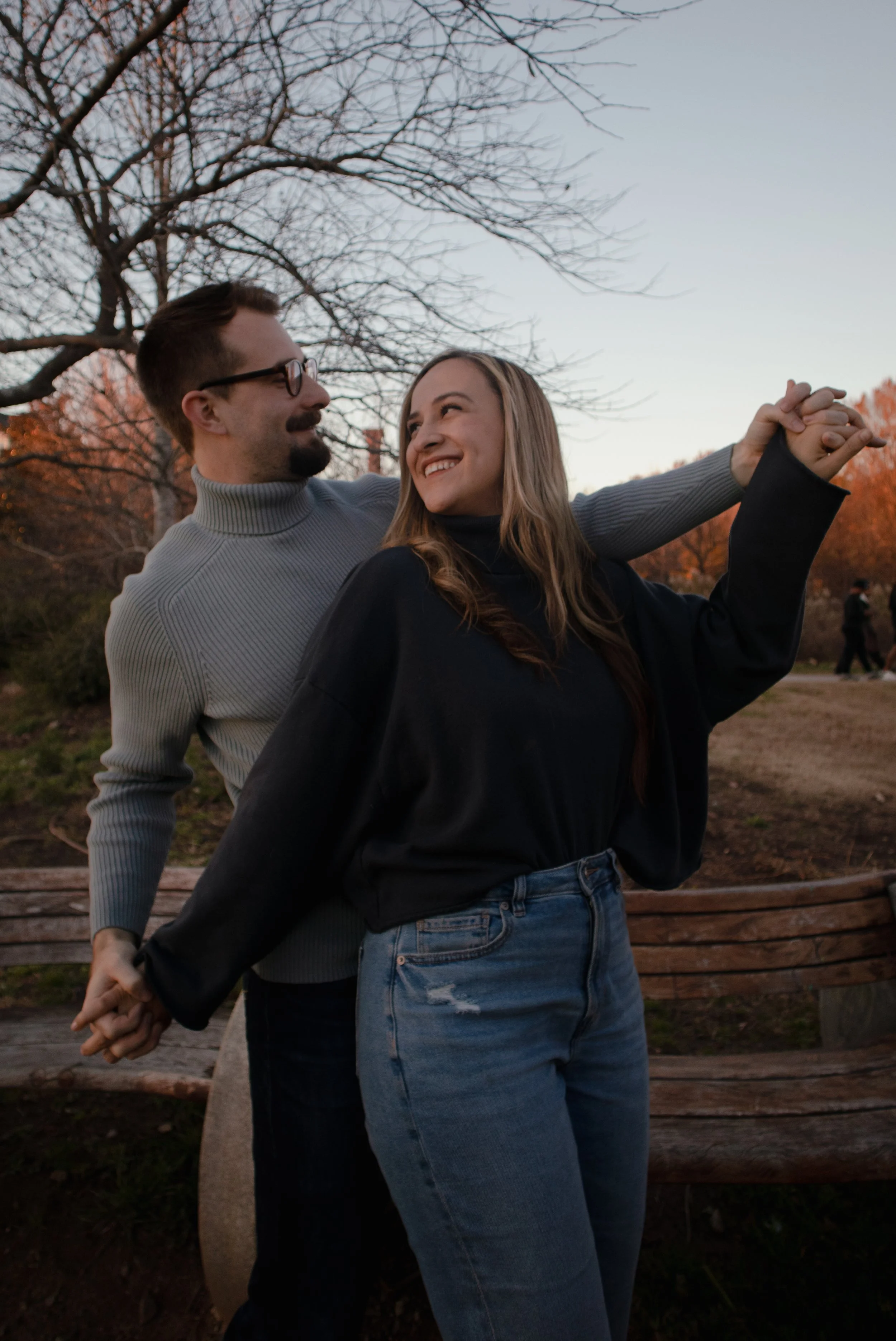 A couple holding hands and dancing outdoors on a park bench during sunset, with trees in the background.