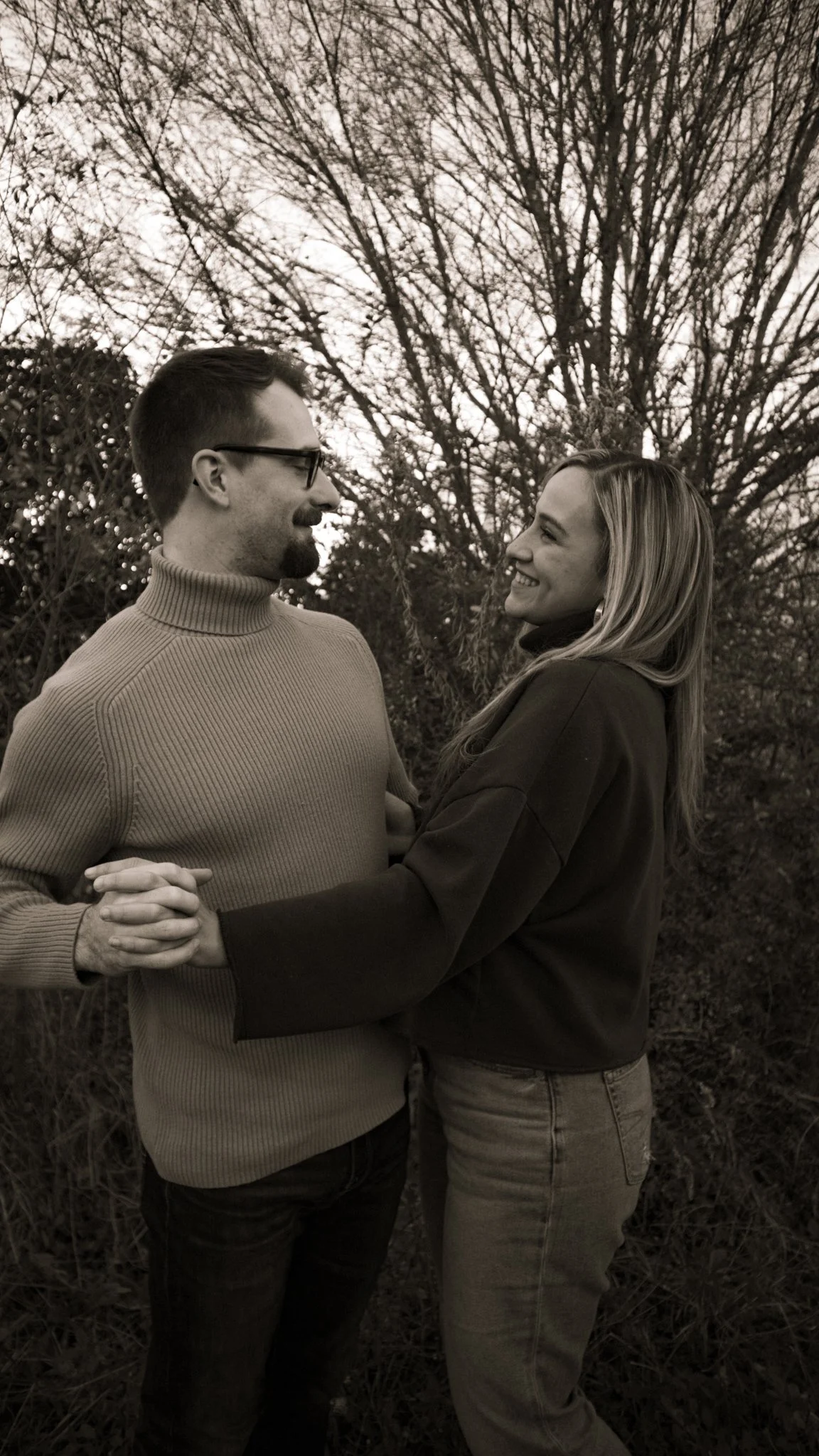 A black-and-white photo of a man and woman facing each other and holding hands outdoors, with leafless trees in the background.