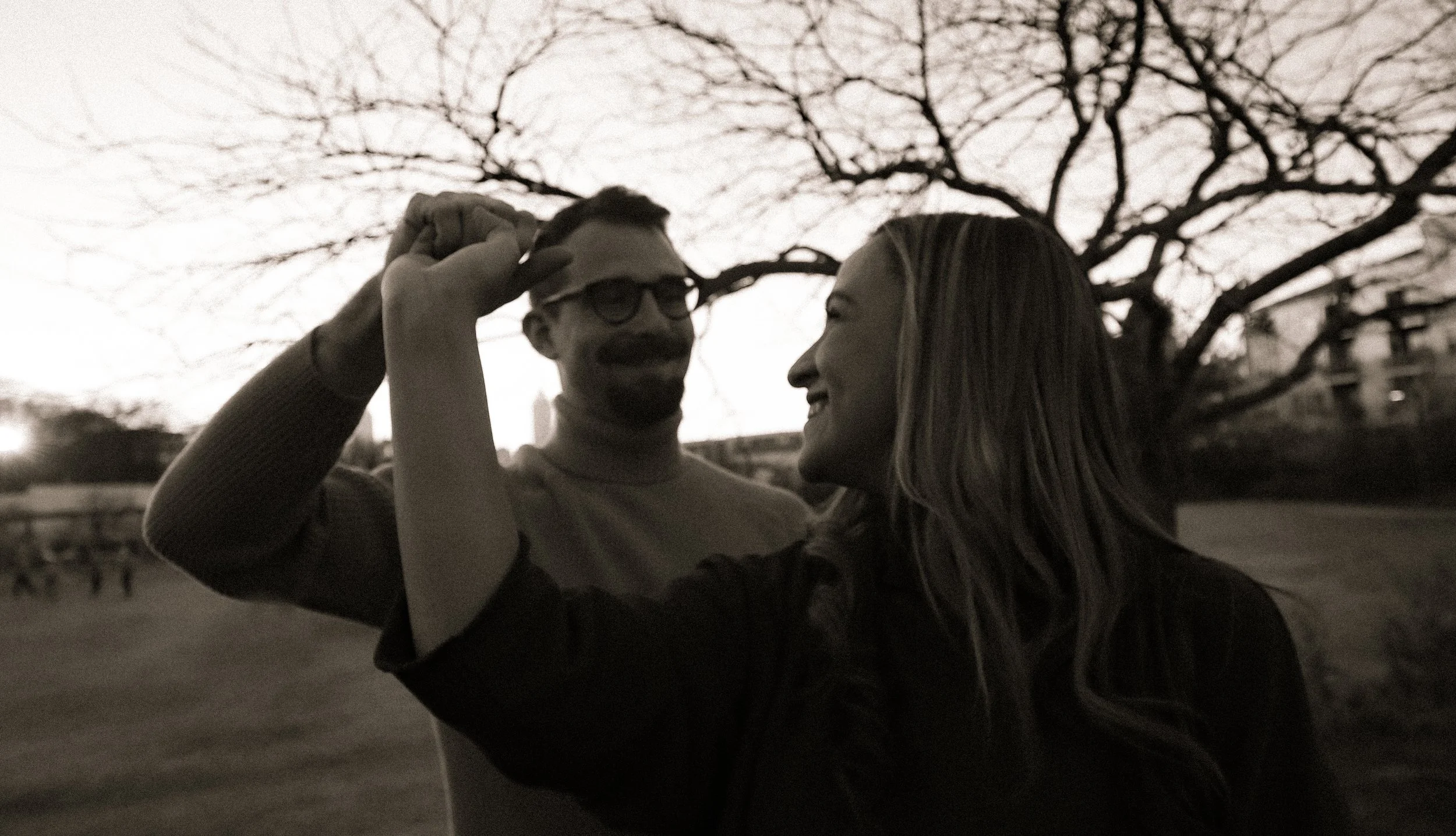 A black and white photo of a couple outdoors, smiling and touching foreheads. The man wears glasses and a turtleneck, while the woman has long hair. A tree with bare branches is in the background.