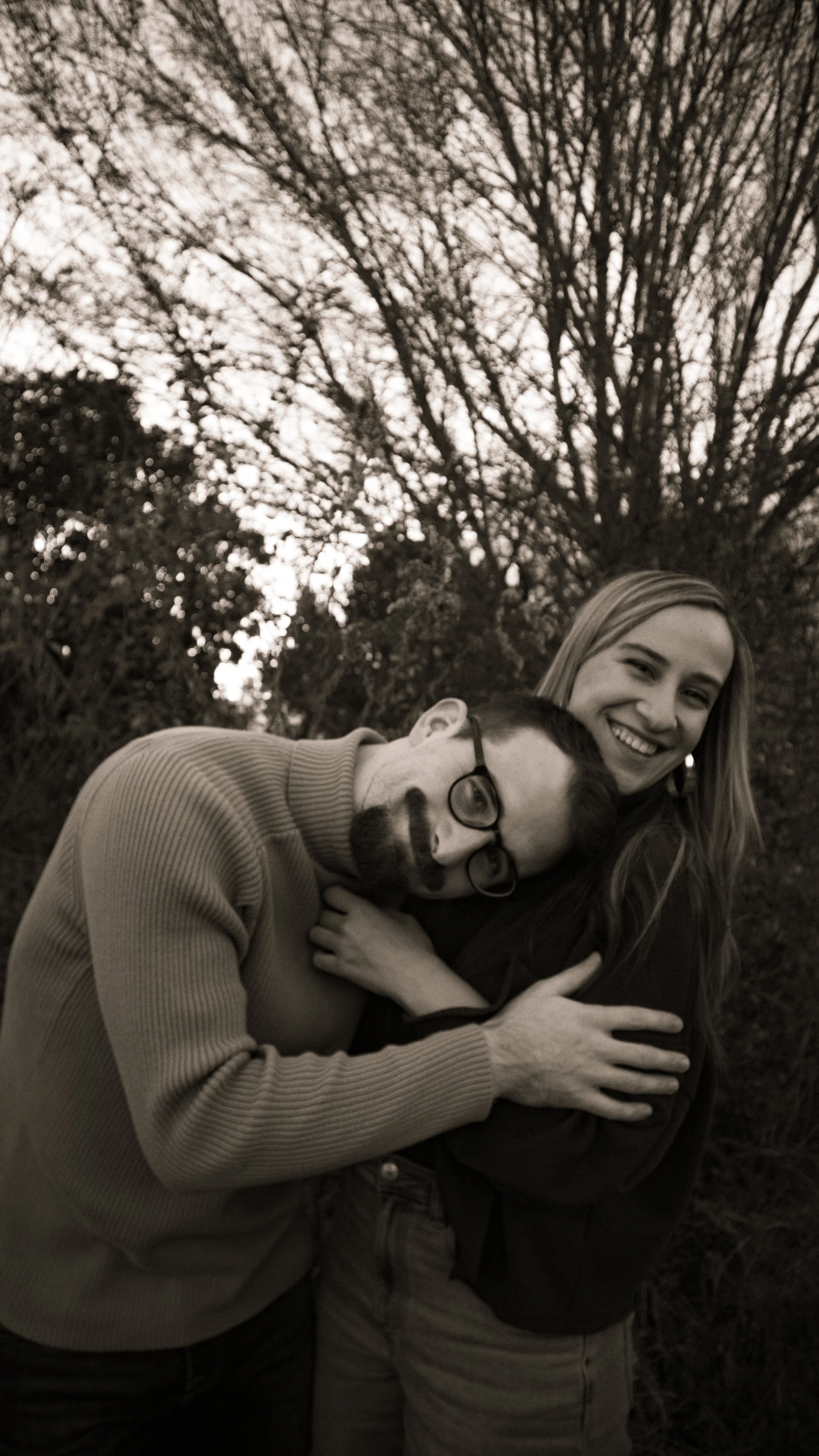 A black and white photo of a smiling woman hugging a man who is wearing glasses and has a beard, in an outdoor setting with trees in the background.