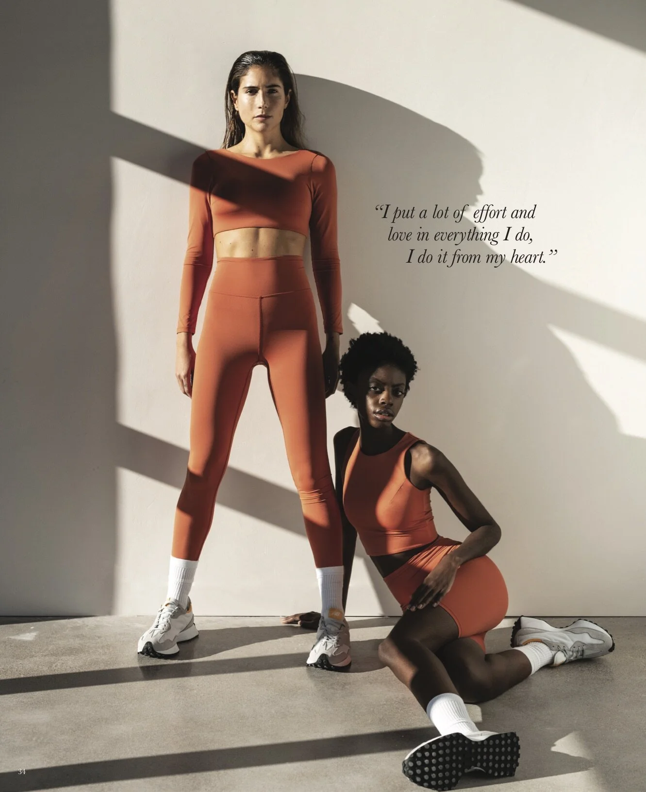 Two women in athletic orange and black workout attire striking poses against a plain wall with shadows. One woman stands while the other is kneeling, with motivational quote on the wall: 'I put a lot of effort and love in everything I do, I do it fro